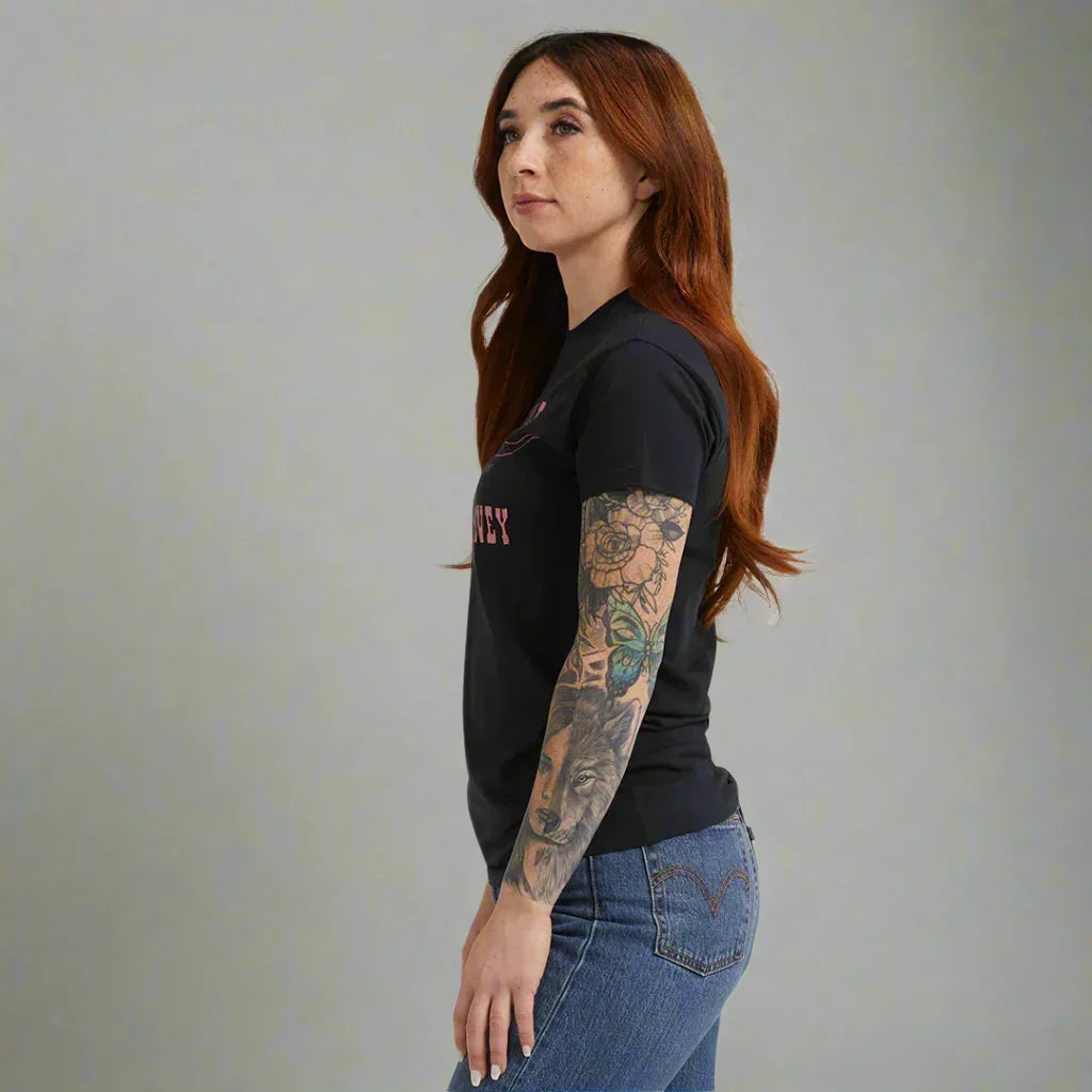 Woman with long red hair in black graphic t-shirt and blue jeans, showcasing colorful arm tattoos