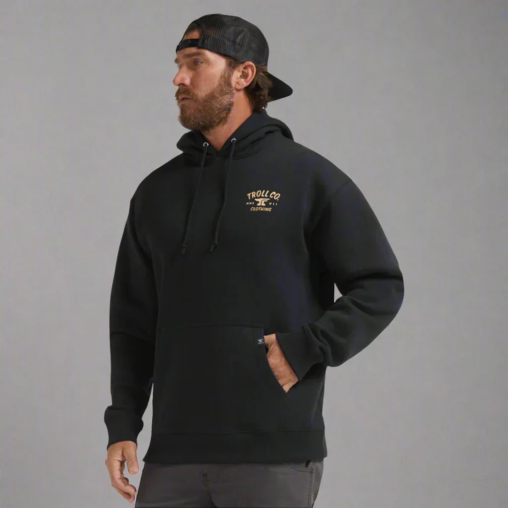 Man wearing black Troll Co. Clothing hoodie and black cap against gray background