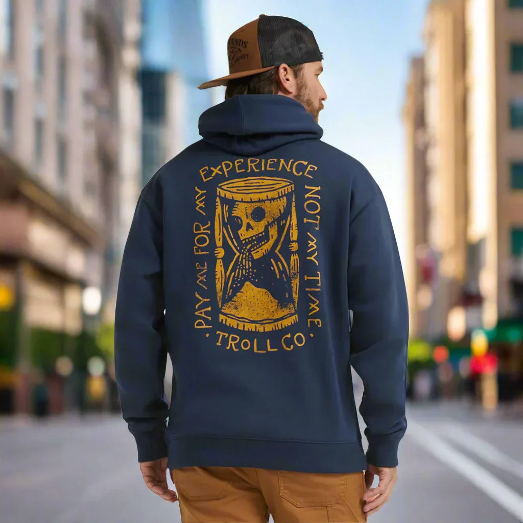 Man in city wearing navy Troll Co hoodie with gold hourglass skull graphic and adventure quote.