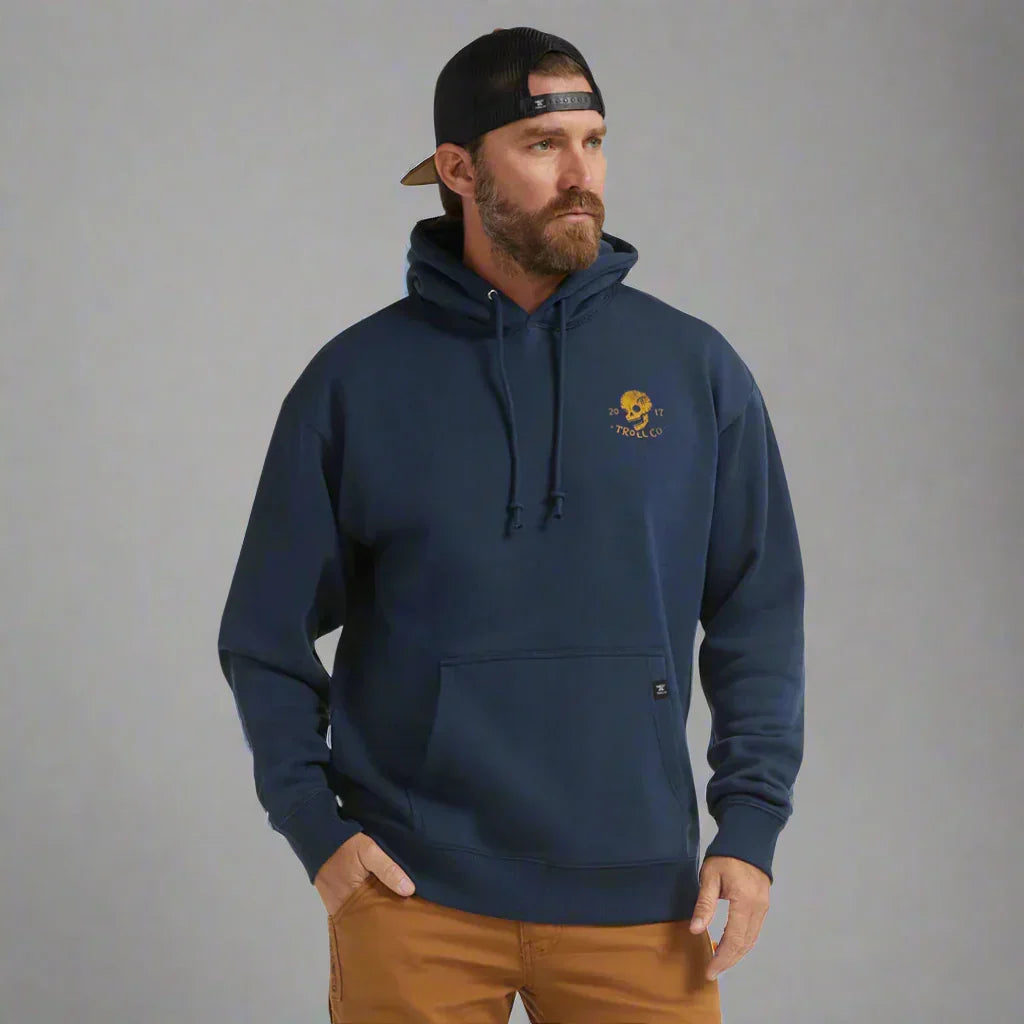 Man in navy blue Troll Co. hoodie and black cap with brown pants against gray background
