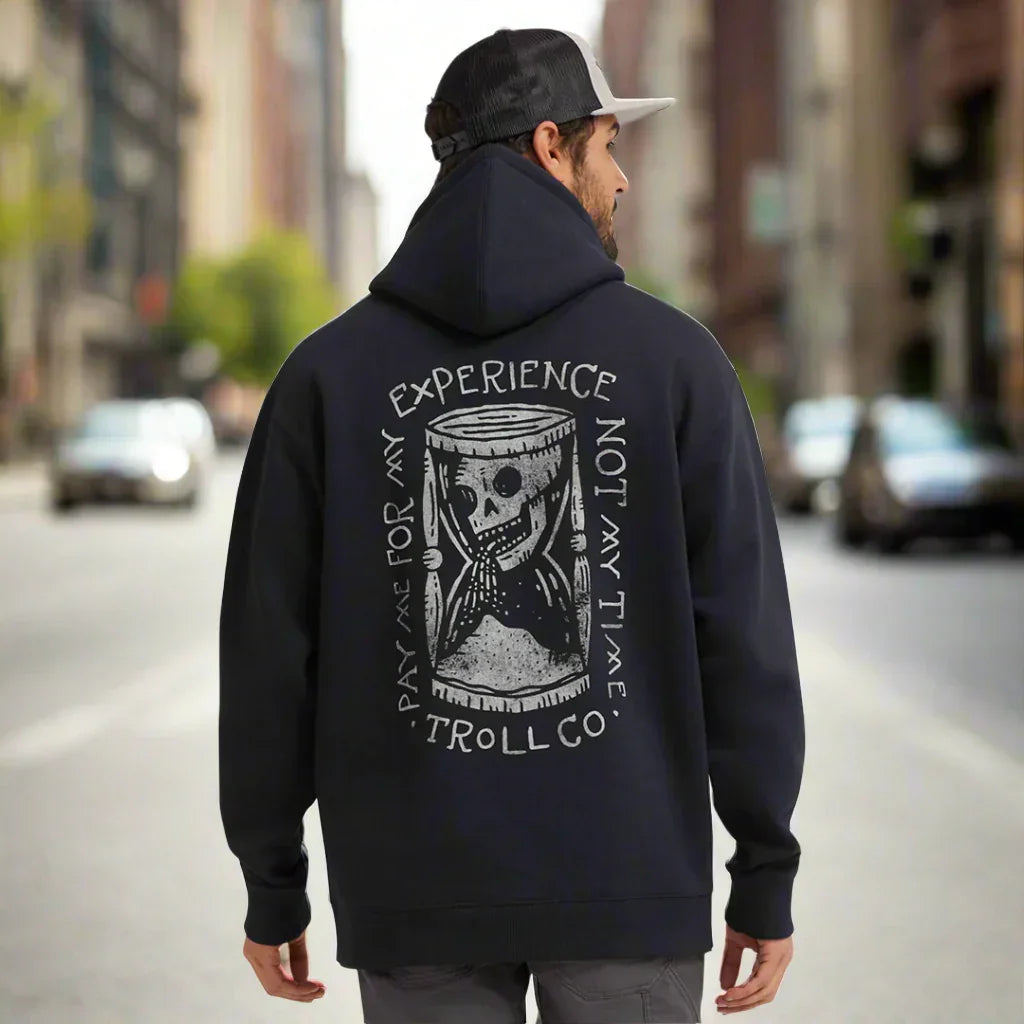 Man wearing black Troll Co. hoodie with hourglass skull graphic and 'Pay me for my experience, not my time' text on city street