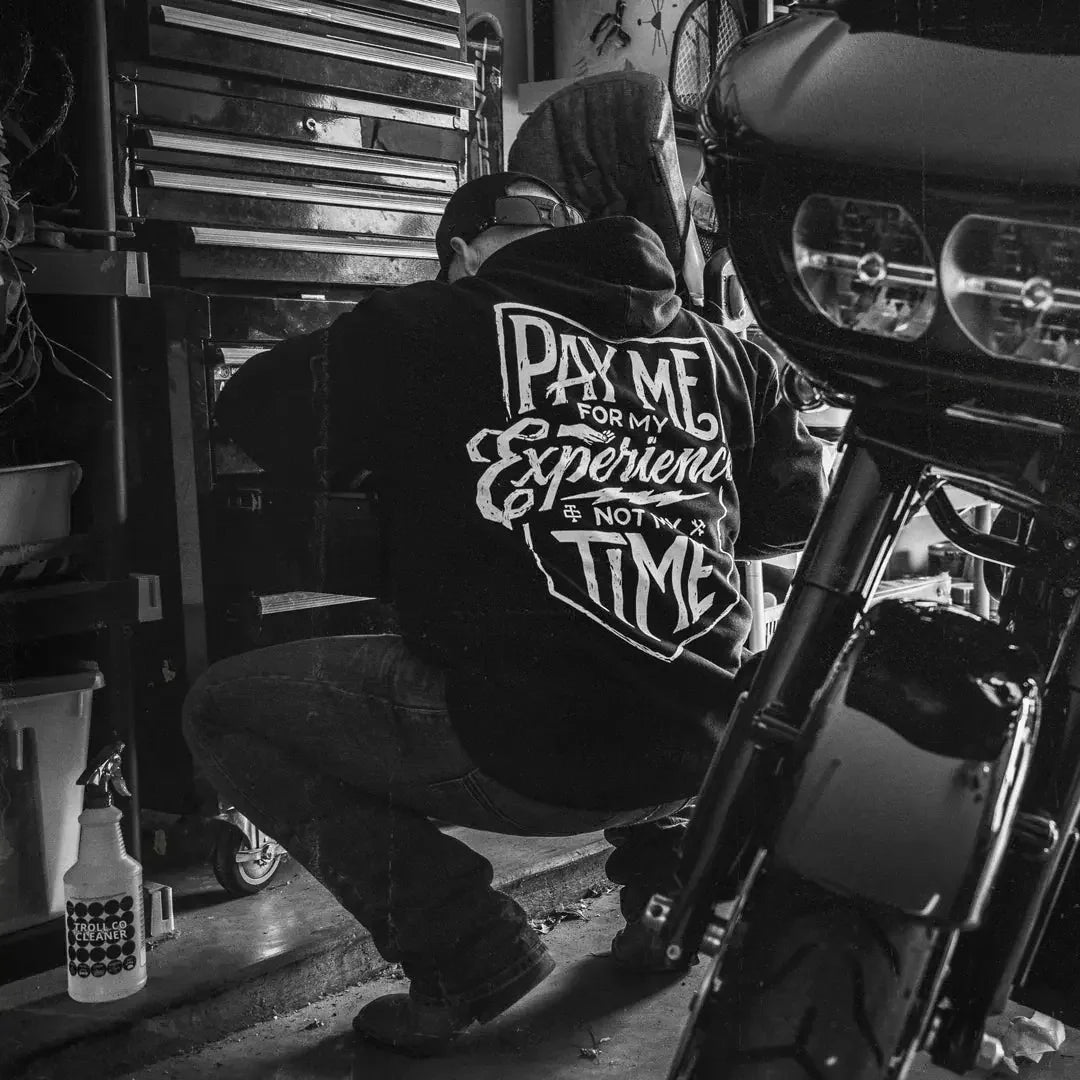Man in Pay Me For My Experience Not My Time hoodie working in garage beside motorcycle.