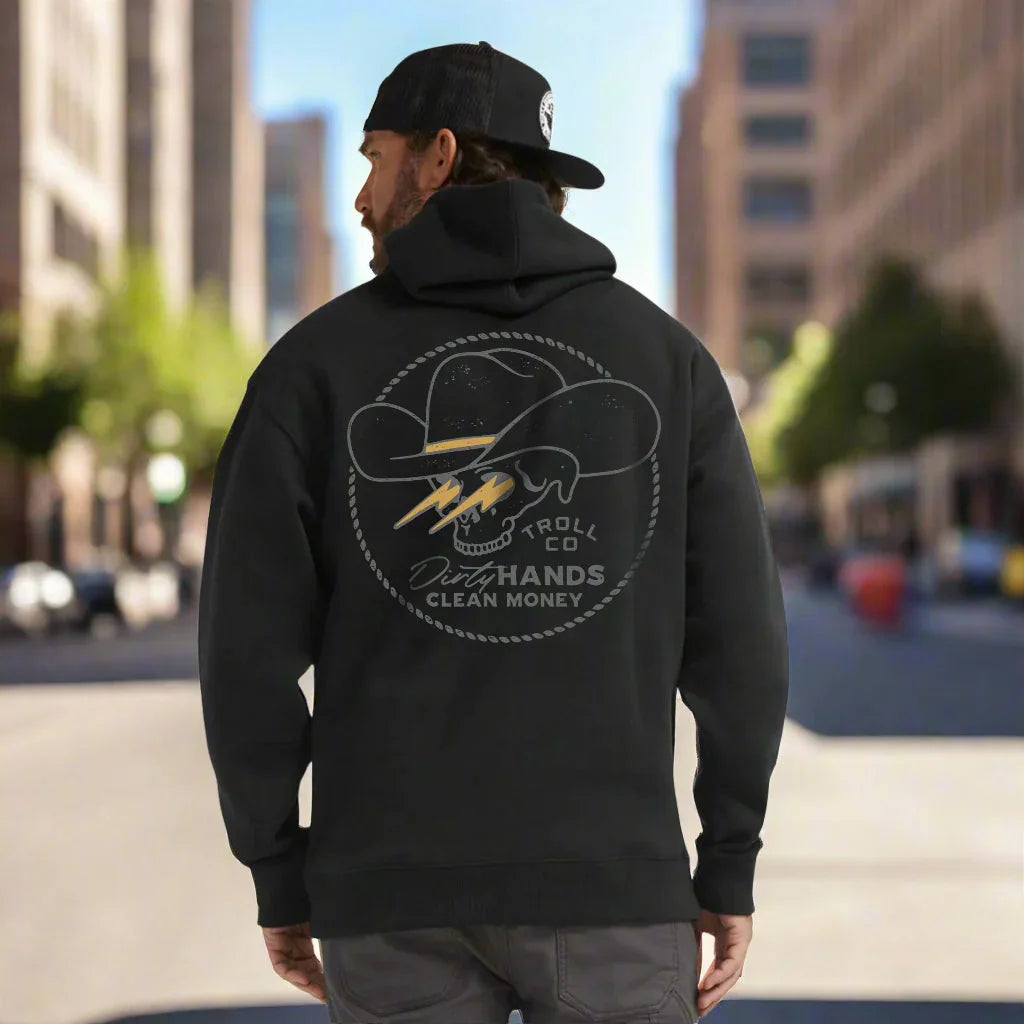 Man wearing black Troll Co hoodie with skull and cowboy hat graphic, urban street background