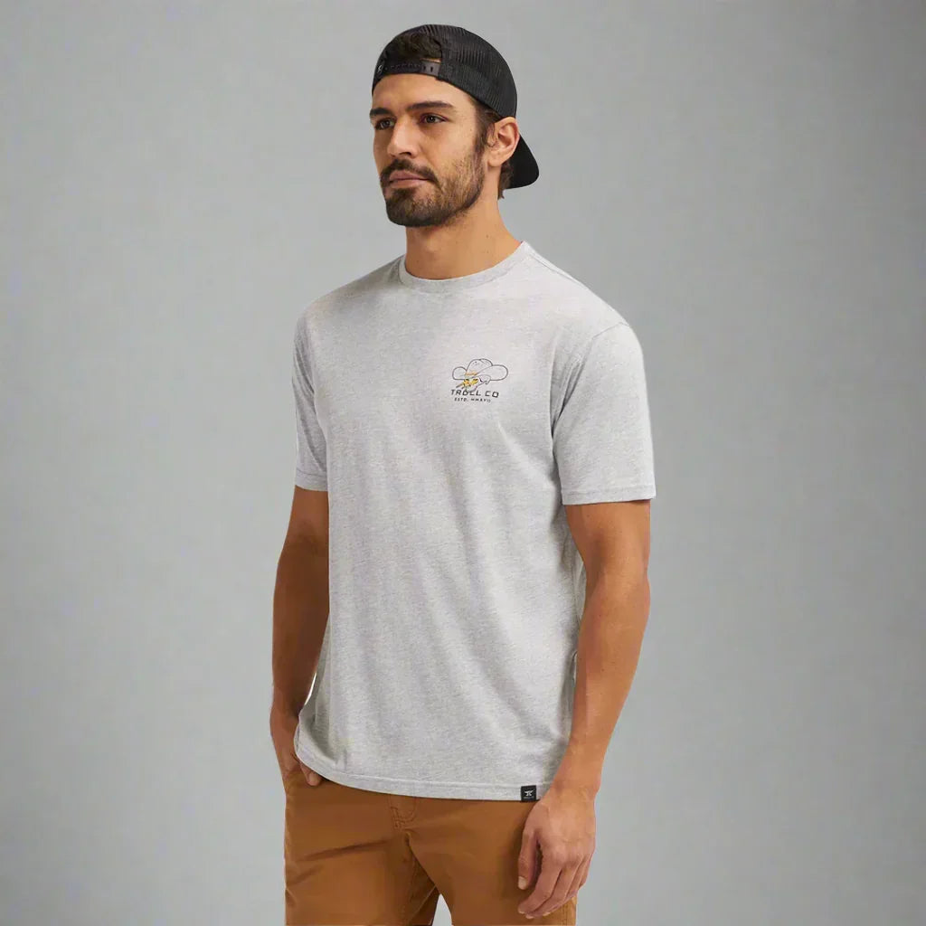 Man wearing light grey Troll Co. t-shirt and black cap, neutral background, casual style