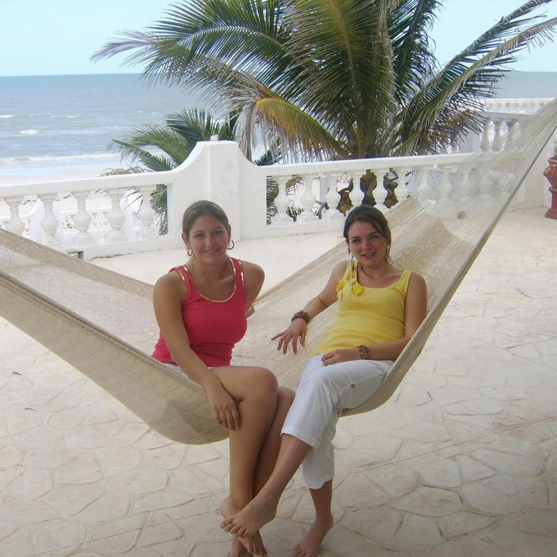 Two women sitting on a Mayan hammock on a beachfront patio with palm trees and ocean view