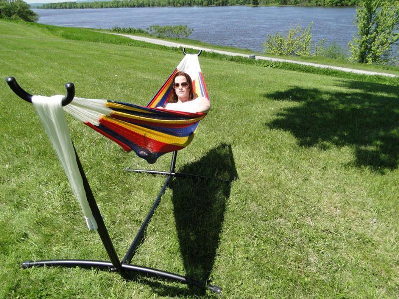 Woman relaxing in colorful Mayan double hammock on universal stand by a river on grass