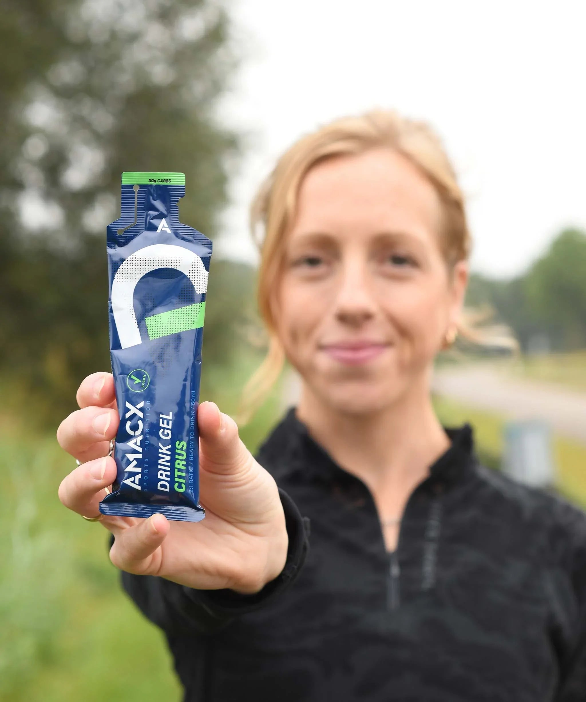Woman outdoors holding Amacx Drink Gel Citrus energy gel with blurred green background