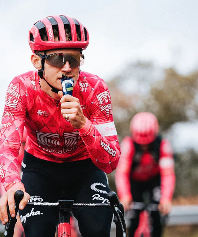 Cyclist in pink EF Education kit using energy gel while riding, wearing helmet and sunglasses