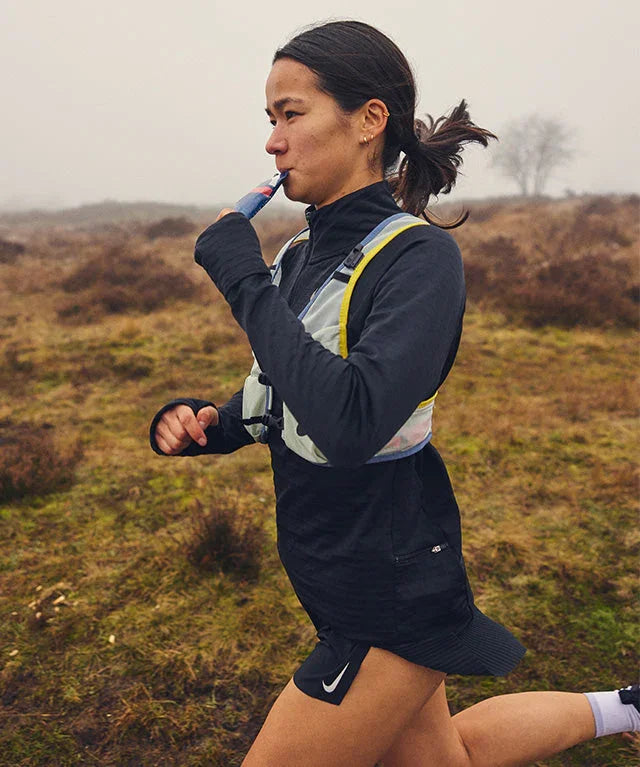 Female runner outdoors drinking strawberry gel, wearing black sportswear and hydration vest