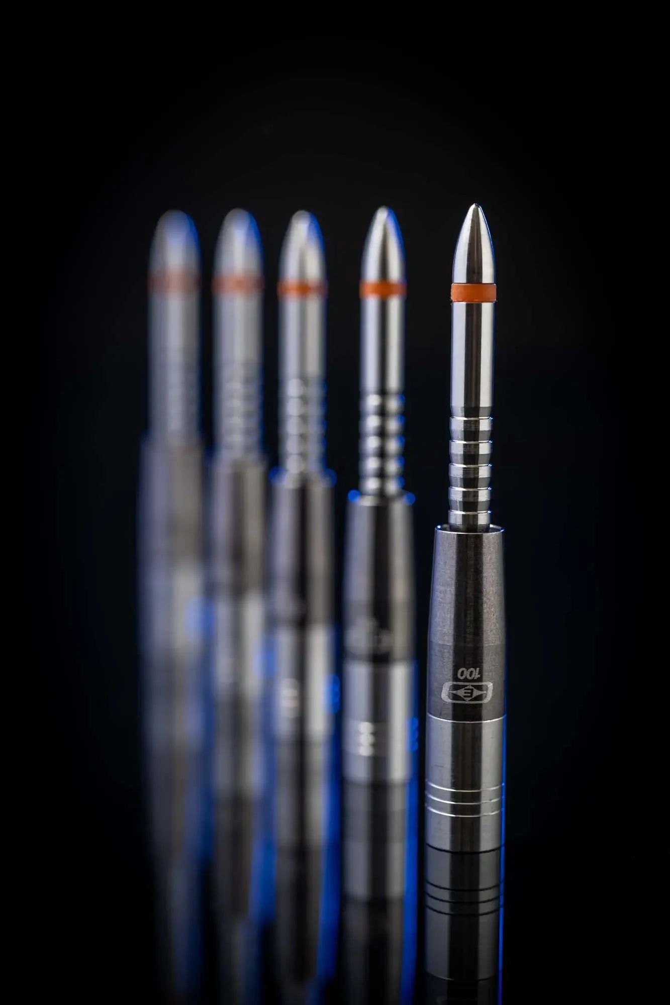 Close-up of five tungsten dart barrels with orange rings, arranged vertically on black background.