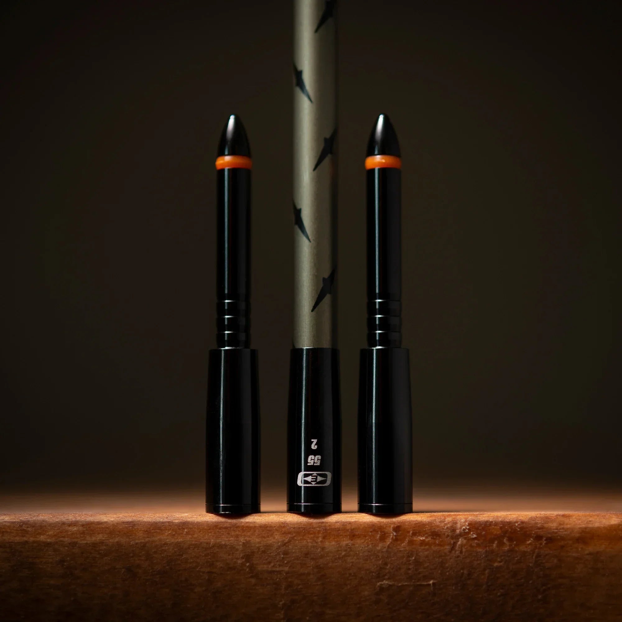 Close-up of two black dart barrels and one dart shaft with orange rings on a brown surface