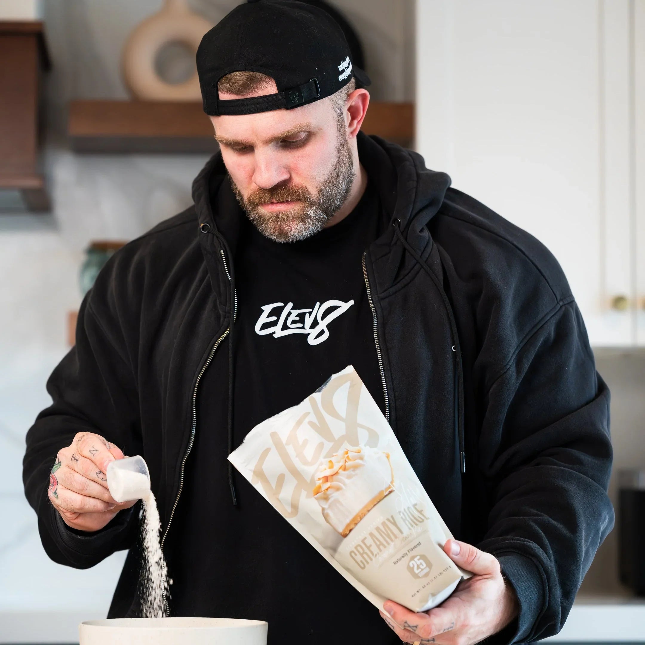 Man in black hoodie using Elev8 Creamy Flake protein powder in modern kitchen