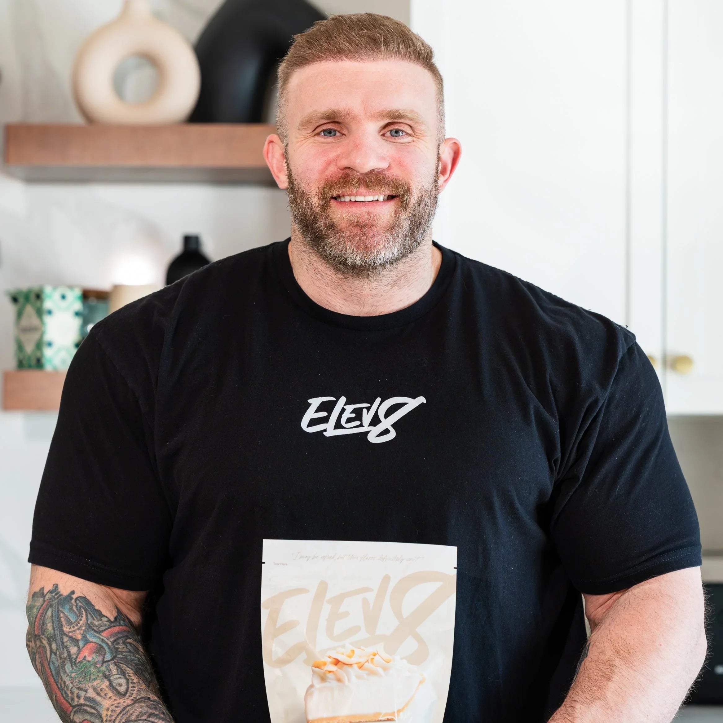 Smiling man with beard in Elev8 shirt holding Elev8 protein bar in modern kitchen