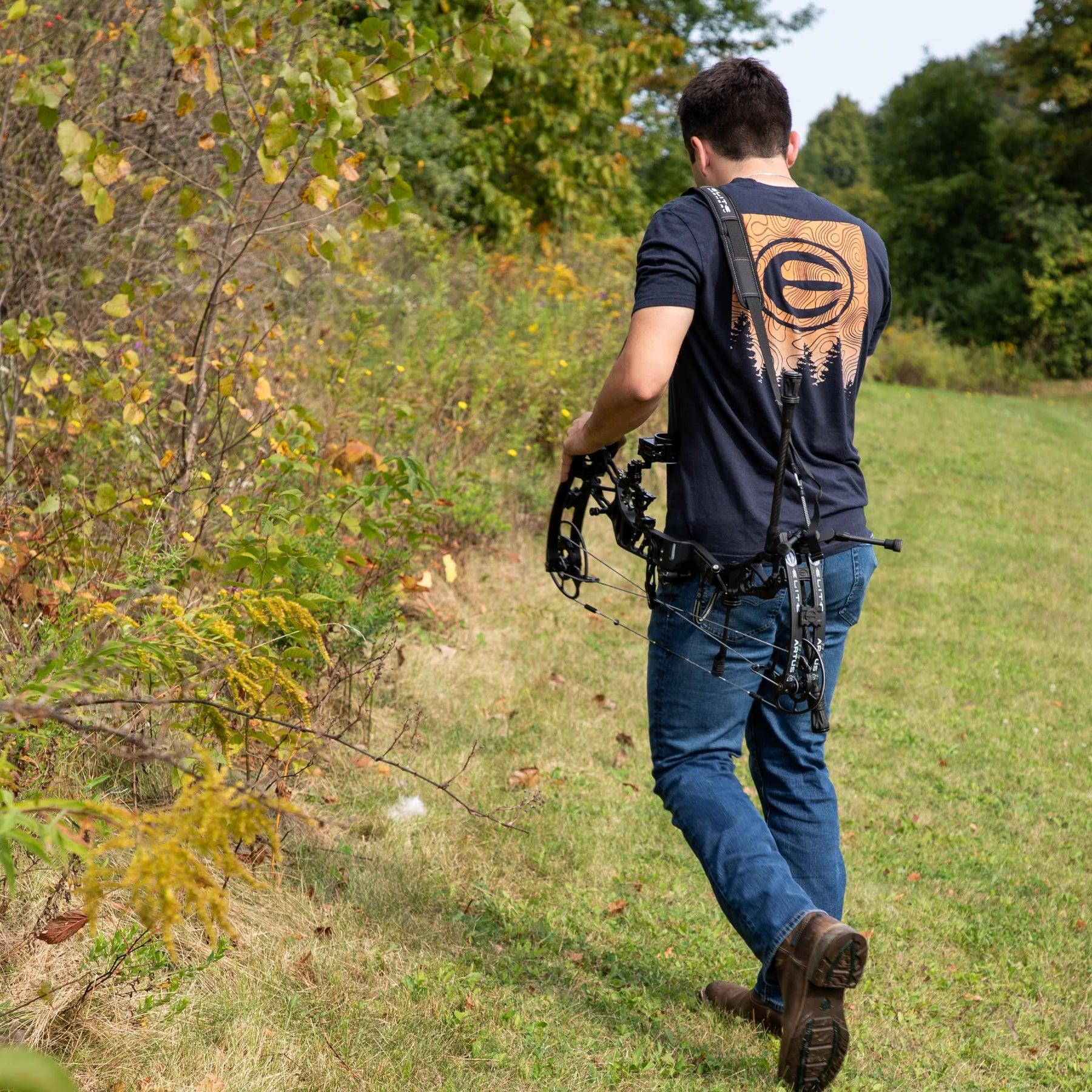 Man outdoors with compound bow and bow sling, walking through grassy field