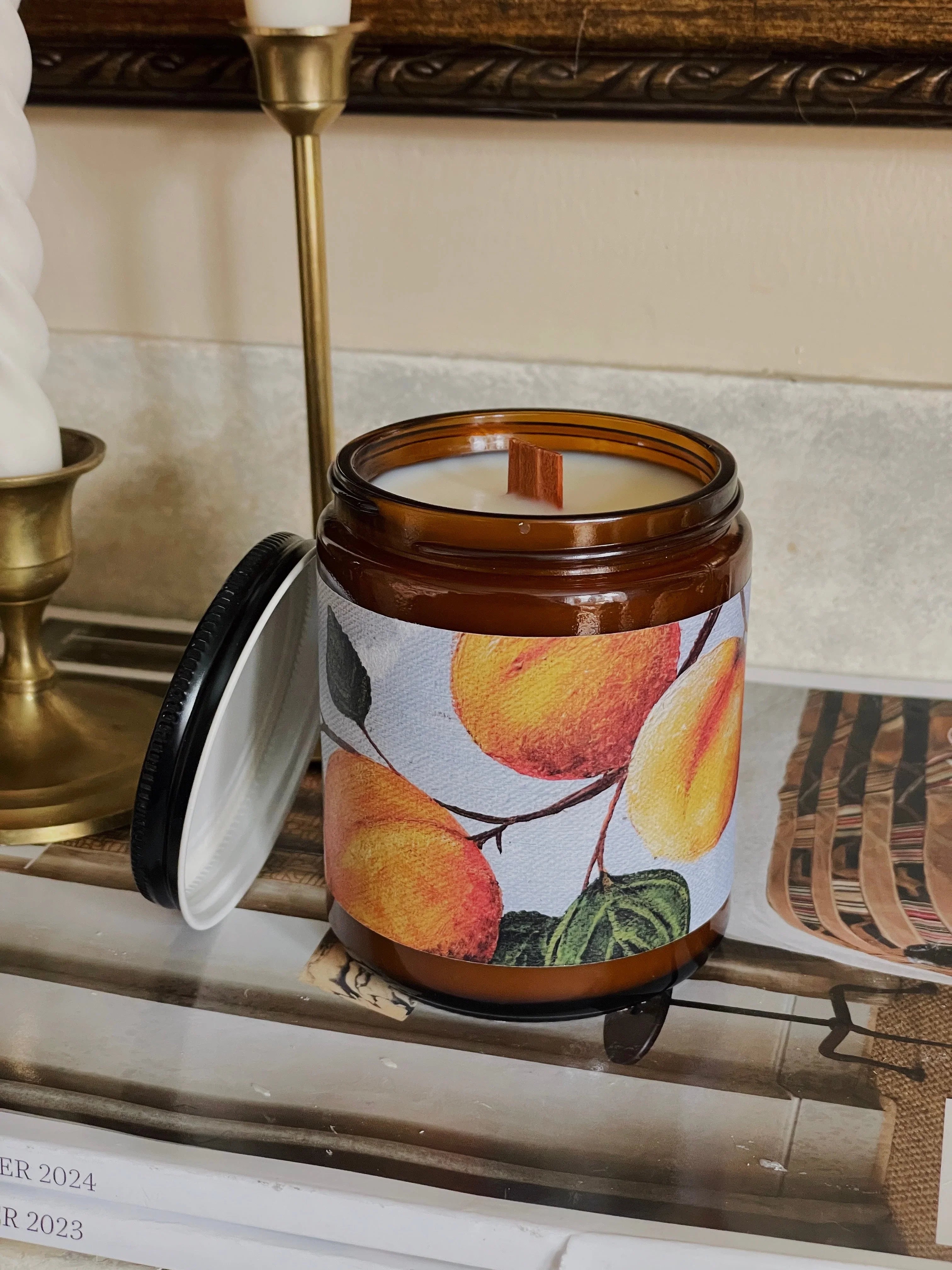 Amber glass candle with fruit label, wood wick, and open lid on stacked magazines