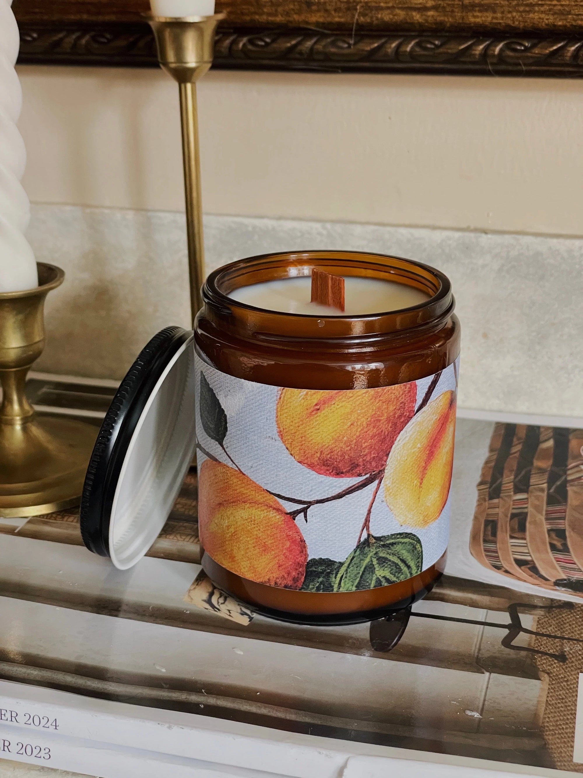Amber glass candle with fruit label, wood wick, and open lid on stacked magazines