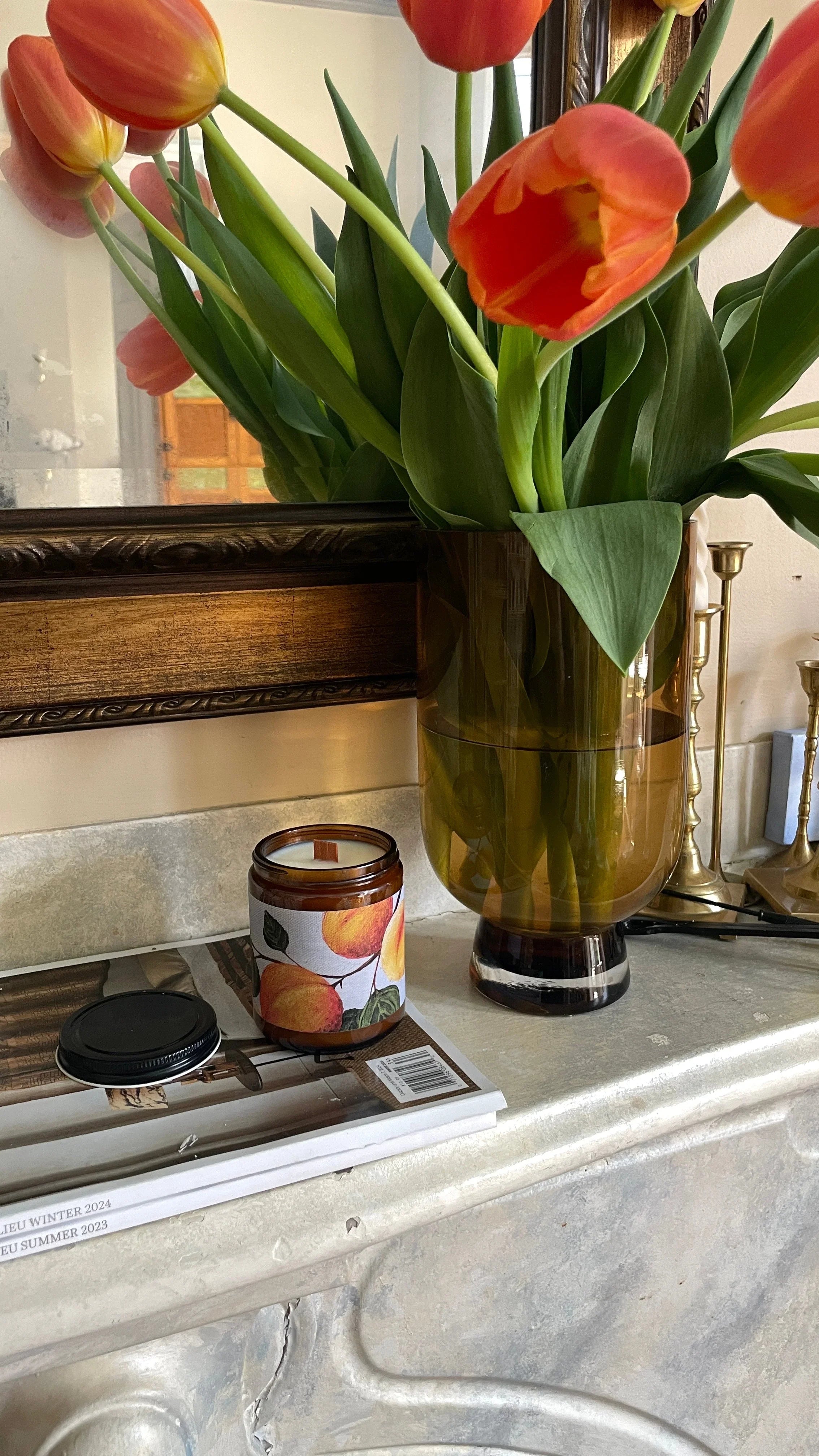 Orange tulips in amber vase, peach-scented candle, and magazines on a marble mantel