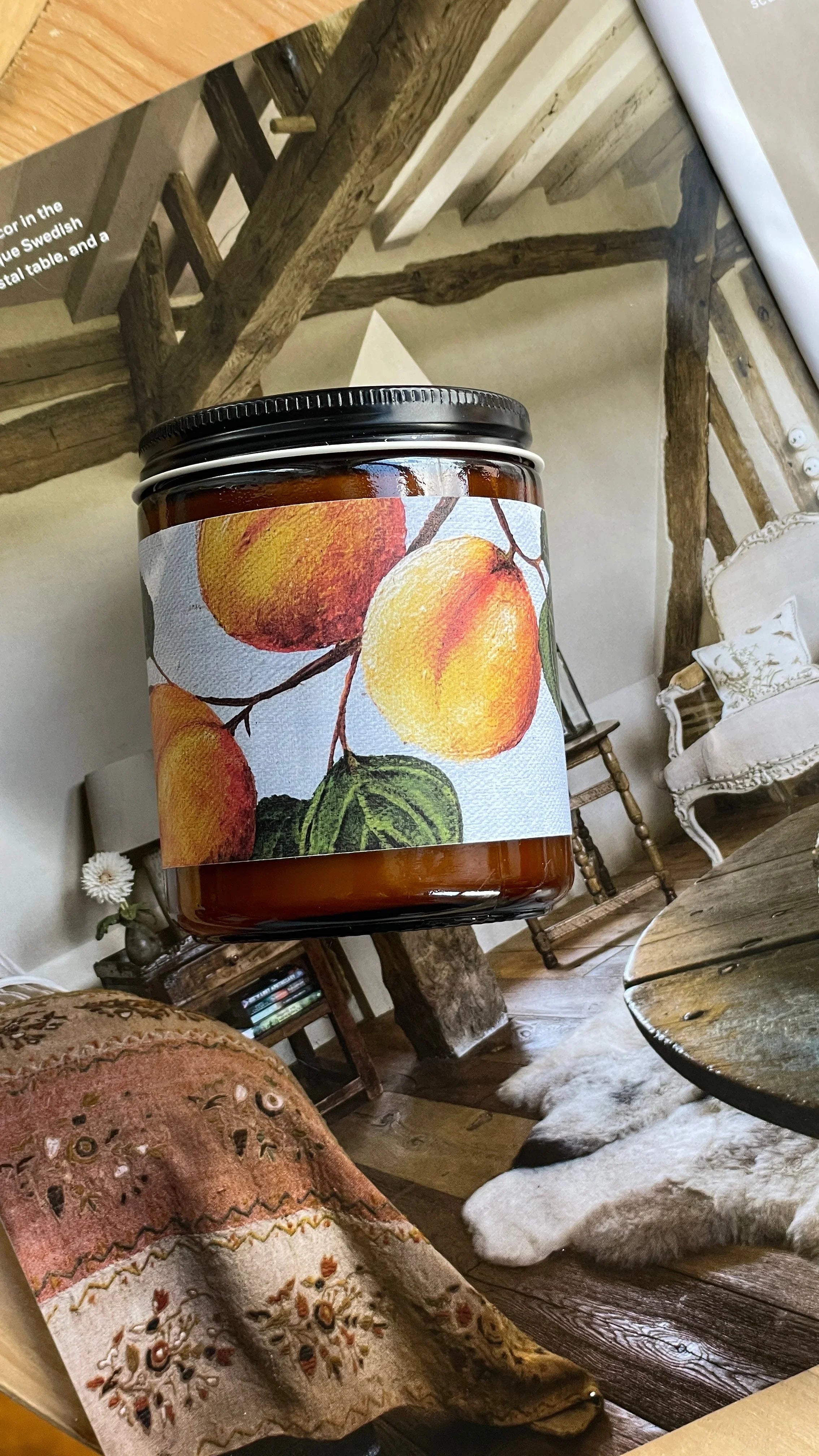 Amber jar candle with peach fruit illustration label, rustic living room background, cozy decor