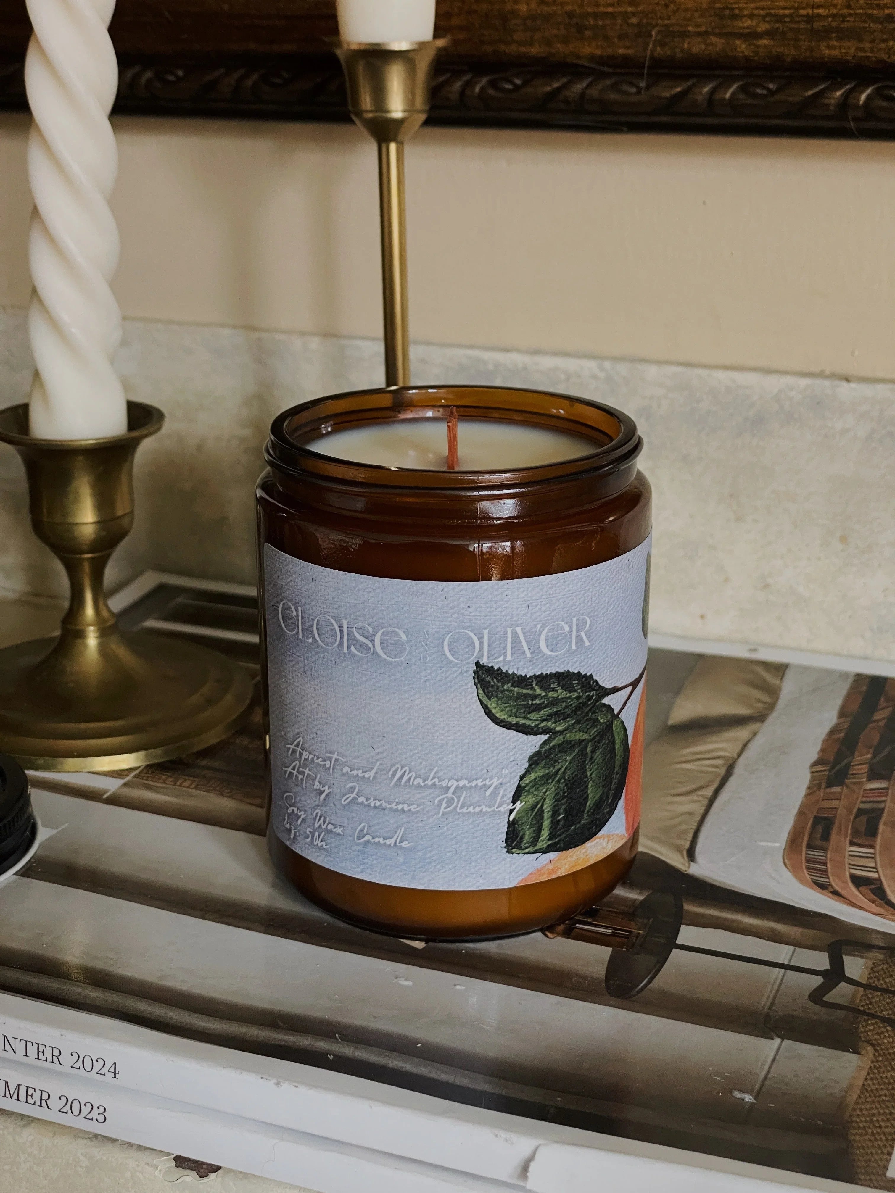 Amber glass Eloise Oliver apricot and mahogany soy candle on books near brass candlesticks