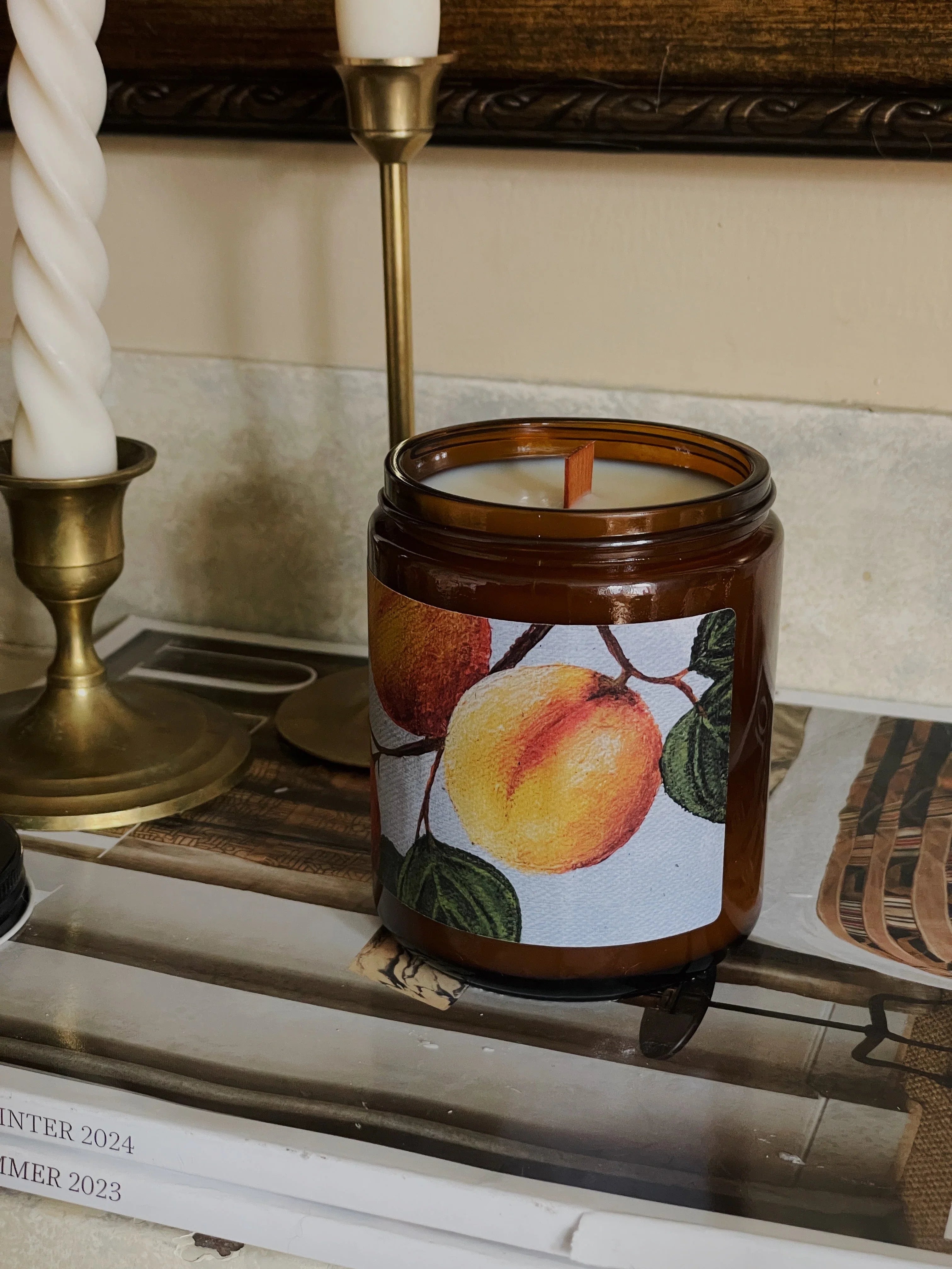 Peach scented candle in amber jar on books with brass candlesticks, cozy home decor