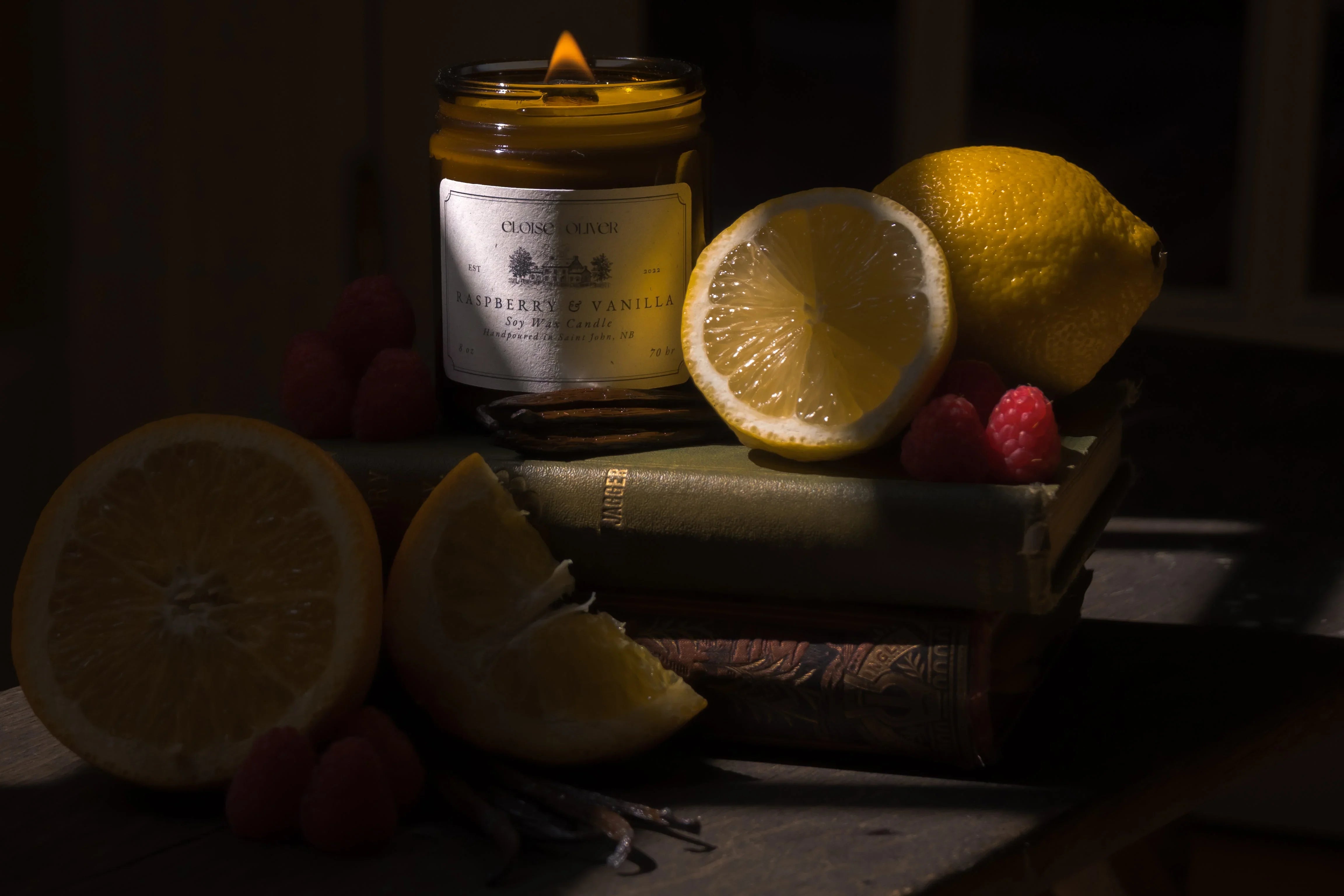 Lit raspberry vanilla soy candle with lemon, orange, raspberries, and books in moody lighting