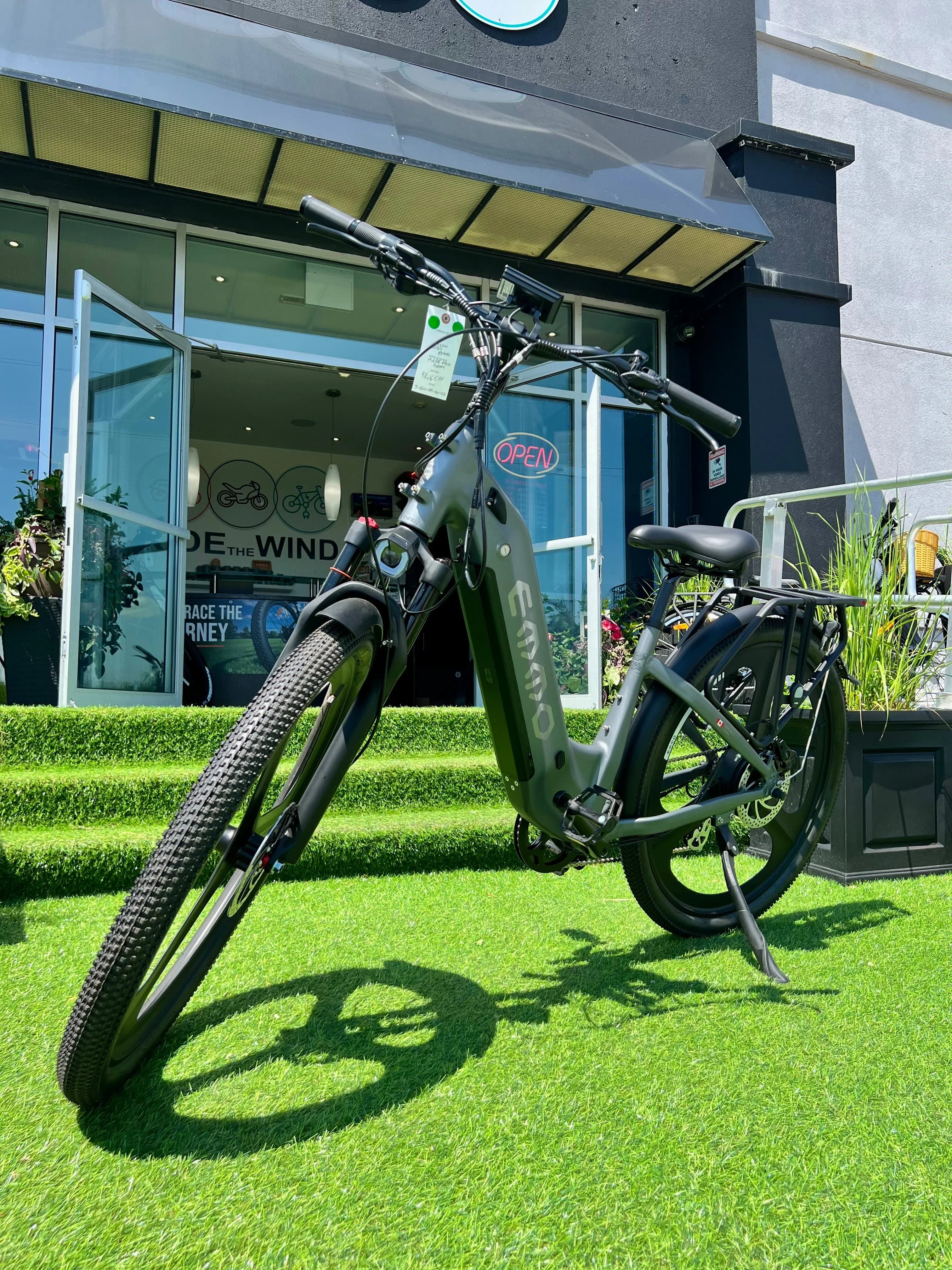 Black EMMO electric bike parked on green artificial grass outside a bike shop with glass doors.