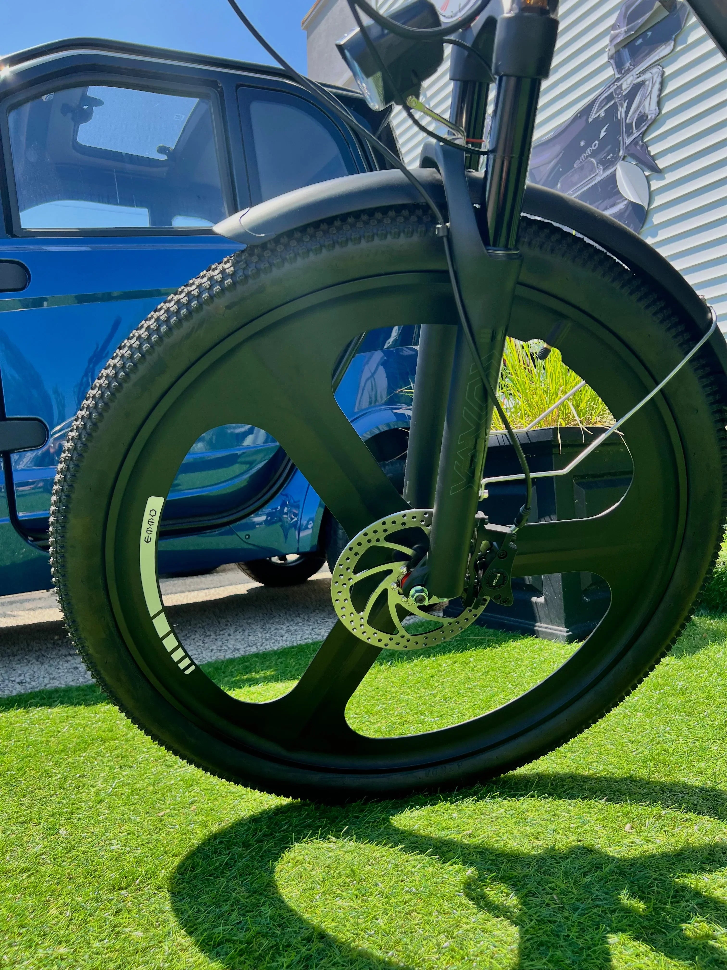 Close-up of black bicycle wheel with disc brake on grass, blue car in background