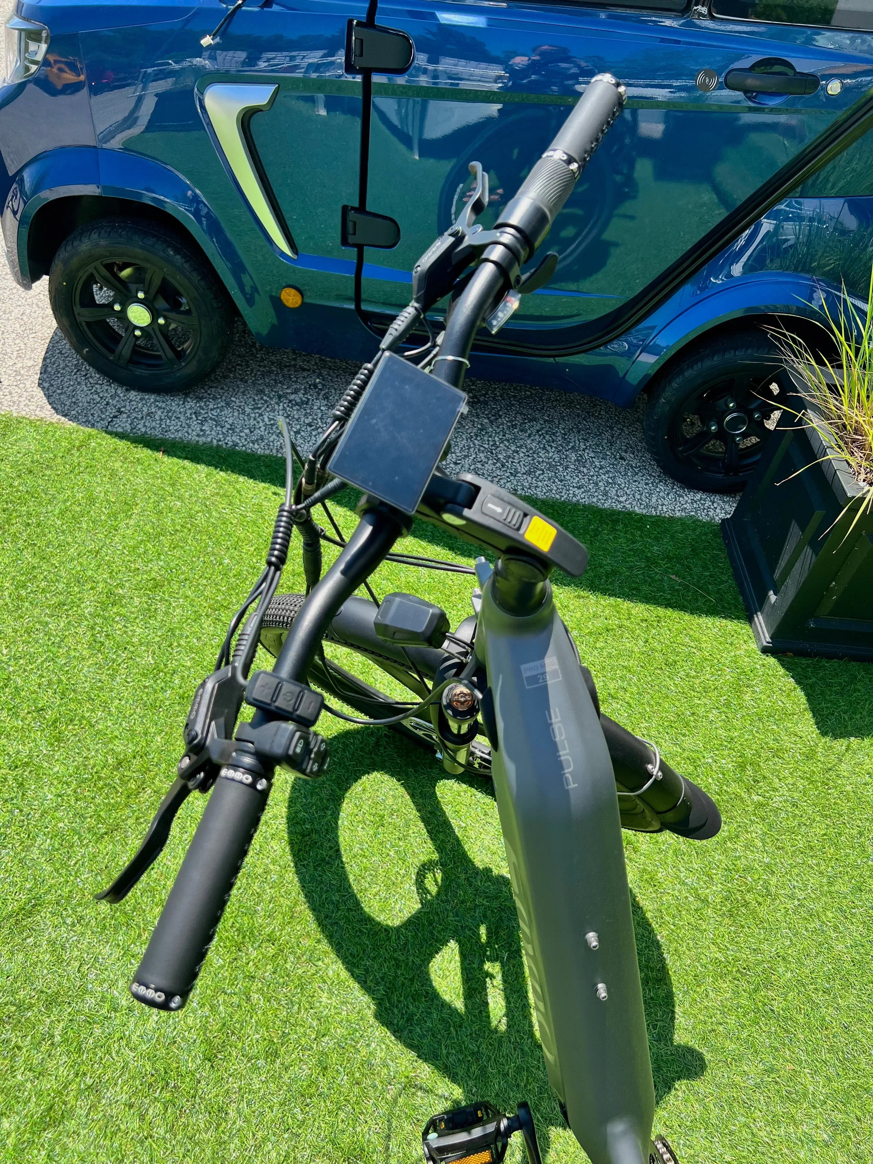 Electric bike handlebars and frame on green grass, blue compact car parked nearby