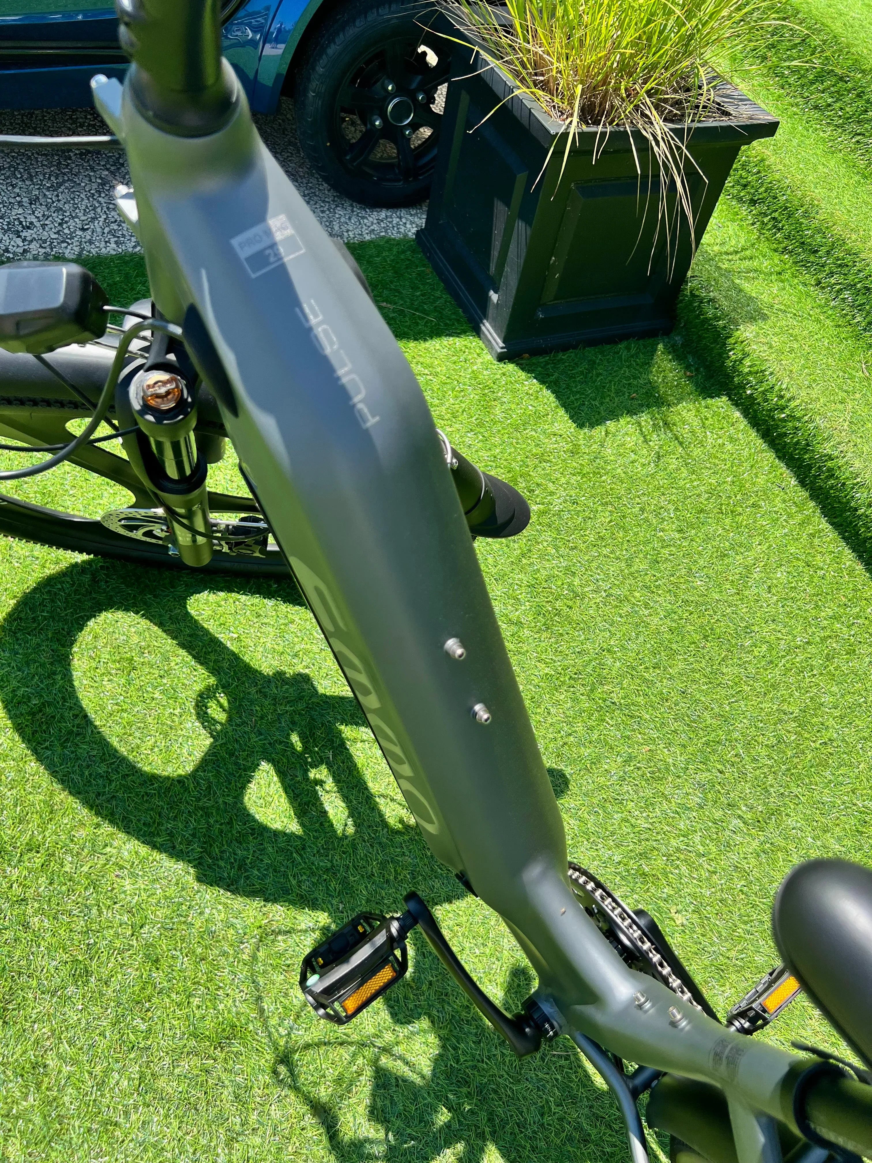 Pulse electric bike frame on green artificial grass outdoors, car and planter nearby