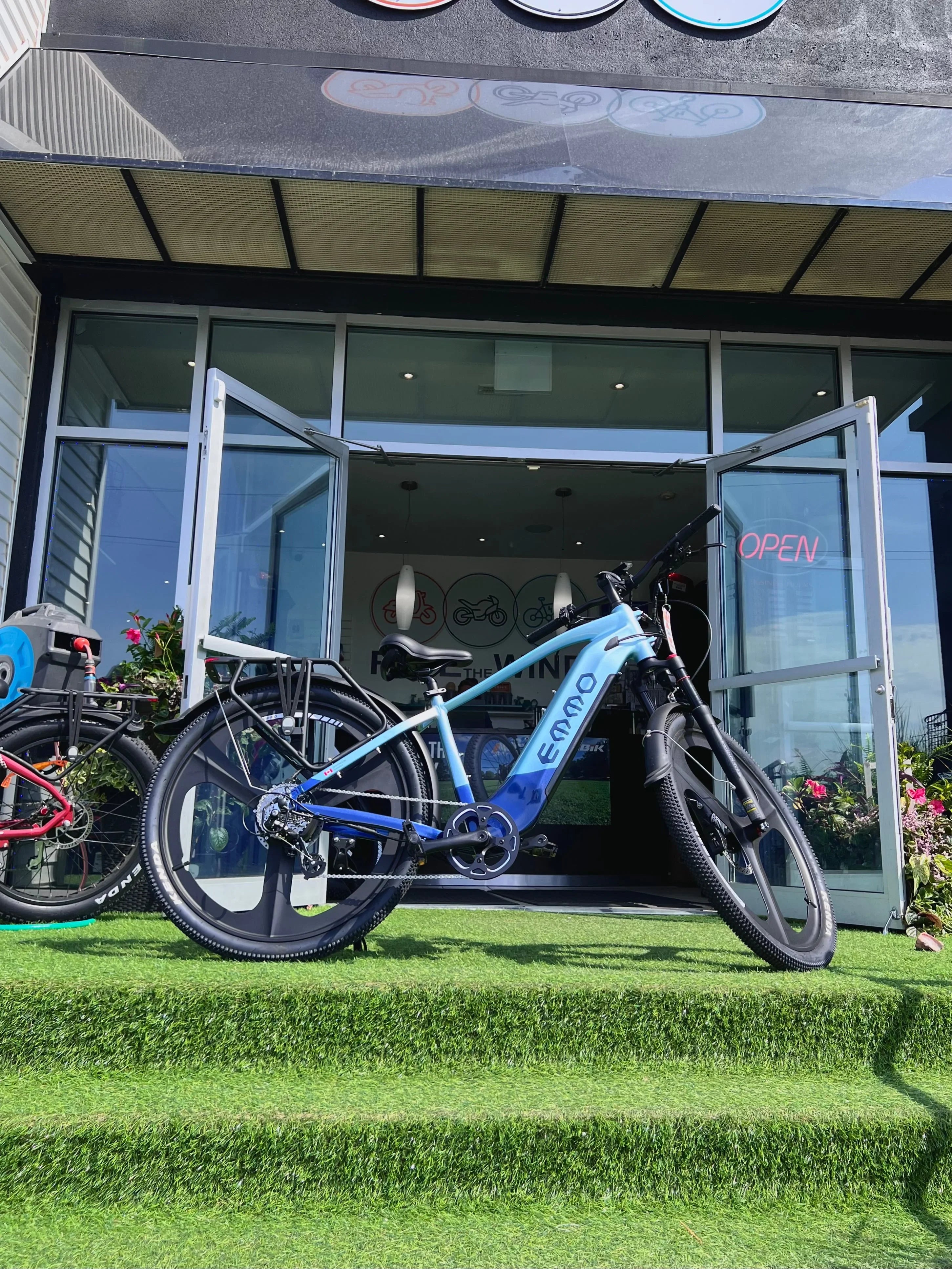 Blue Emmo electric bike displayed outside bike shop with open glass doors and grass steps
