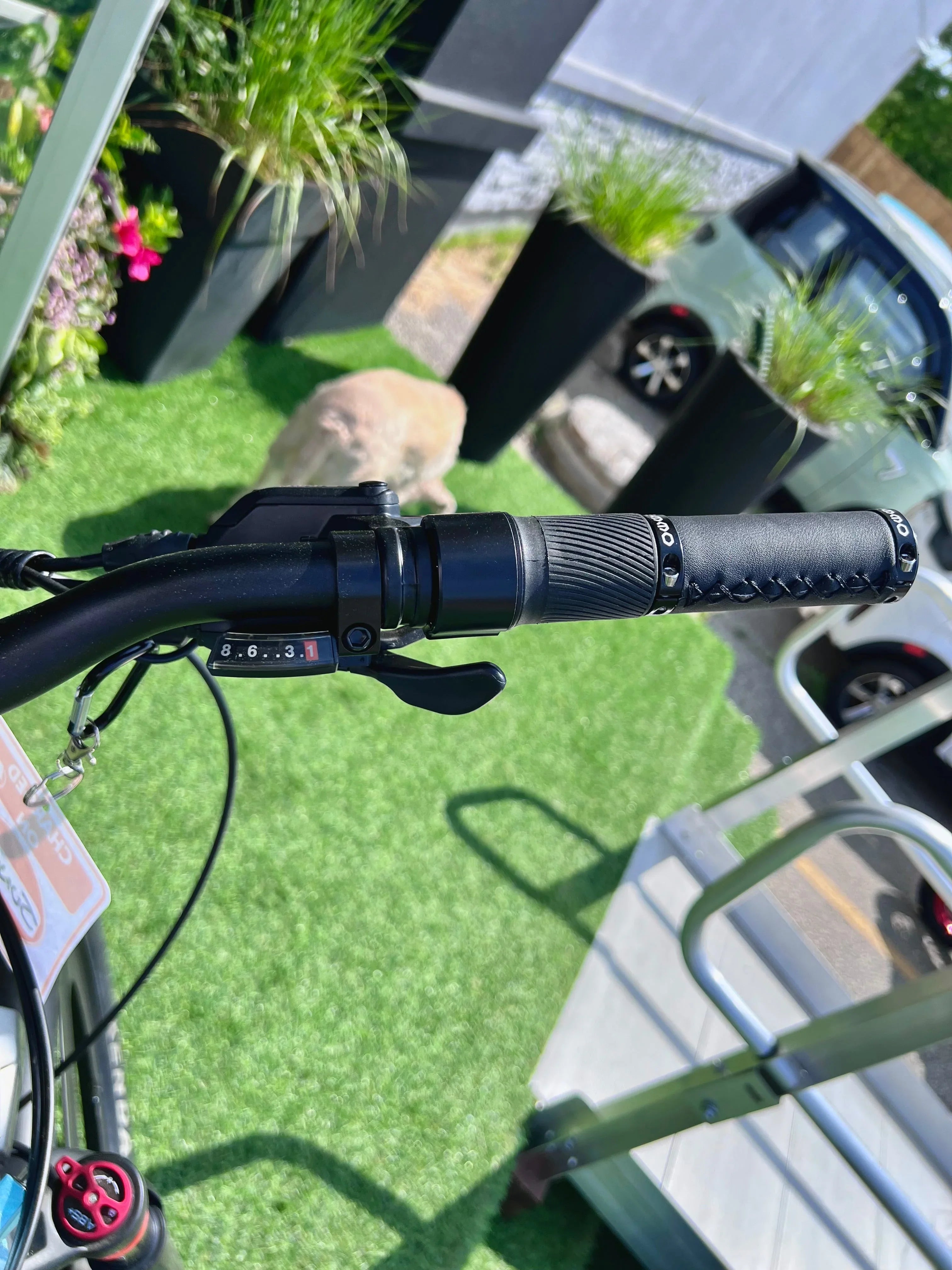 Bicycle handlebar with gear shifter, green artificial grass, planters, and parked car