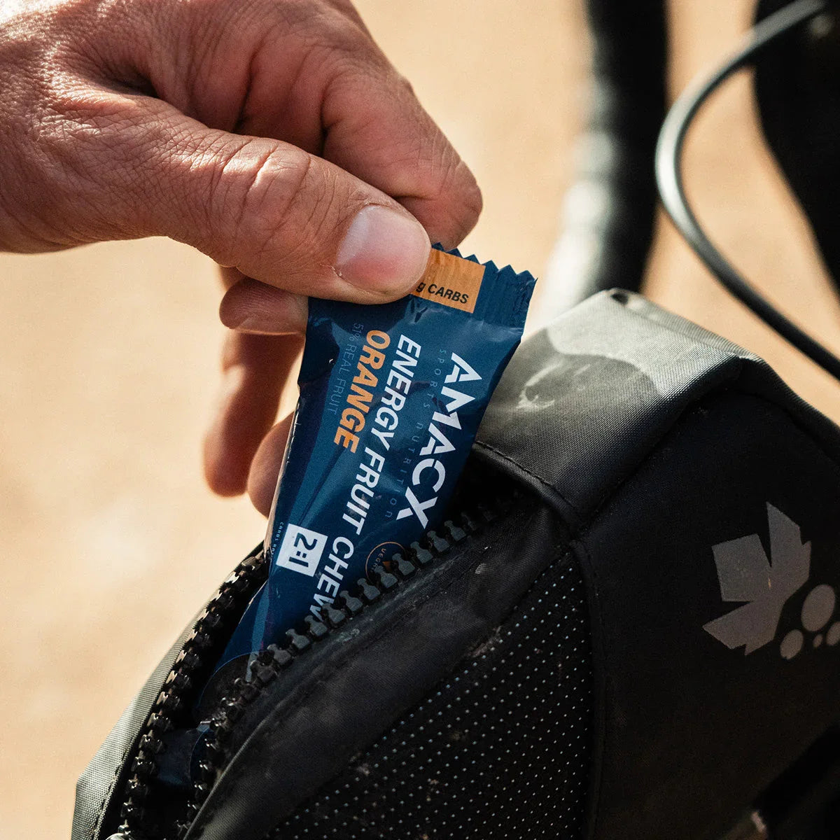 Hand placing Amacx Energy Fruit Chew Orange bar into black bike bag outdoors