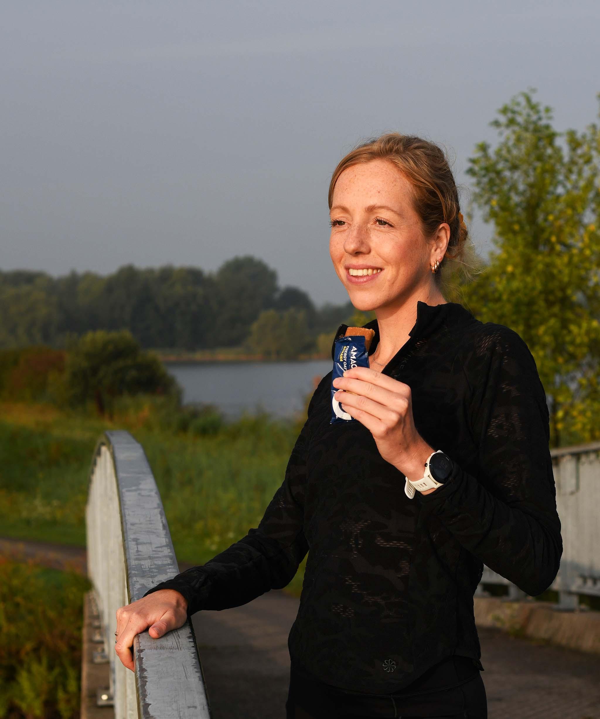 Woman runner eating Energy Oat Bar banana on outdoor bridge near nature and water