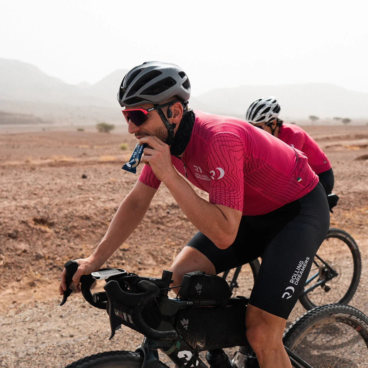 Cyclist in pink jersey and helmet eats oat bar while riding bike in desert landscape