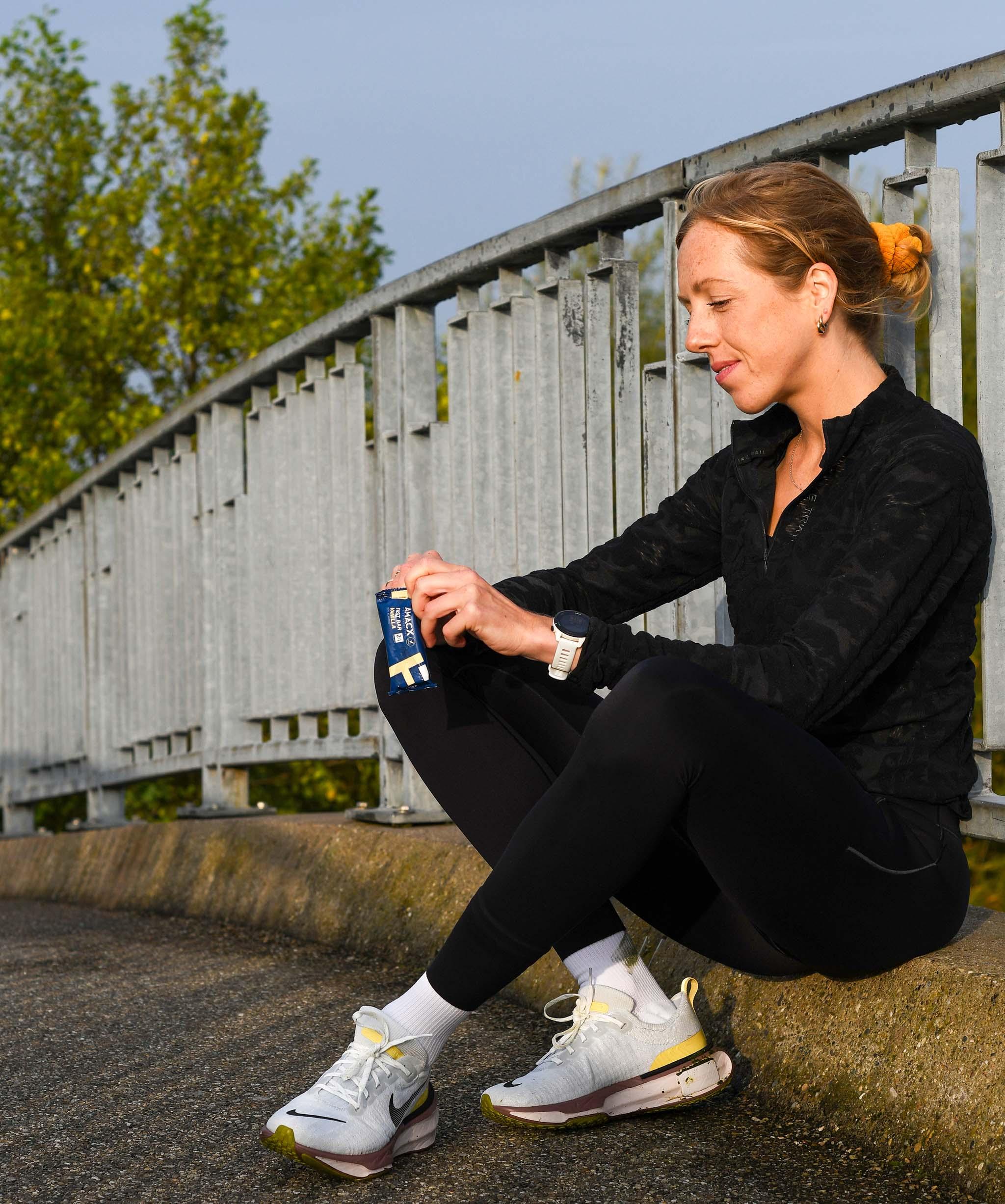 Woman in athletic wear sitting outdoors on a bridge, holding FastBar Vanilla nutrition bar