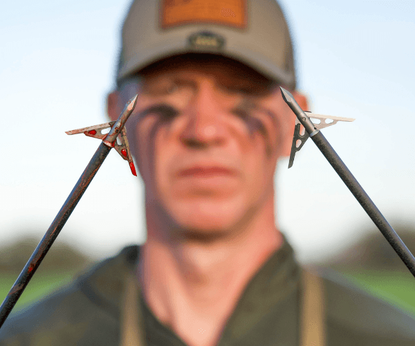 Hunter with camo face paint holding two broadhead arrows outdoors