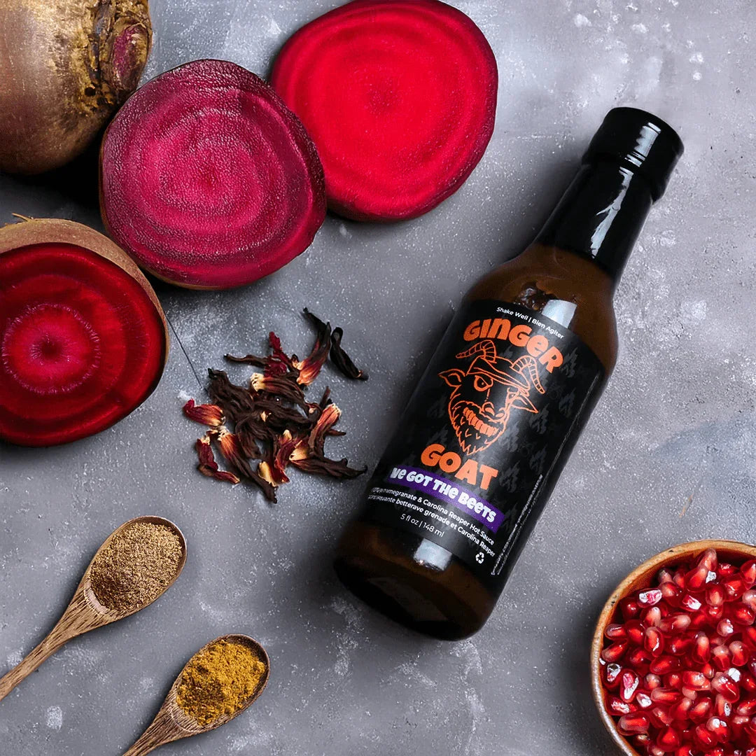 Ginger Goat beet hot sauce bottle with sliced beets, pomegranate seeds, spices, and hibiscus on gray surface