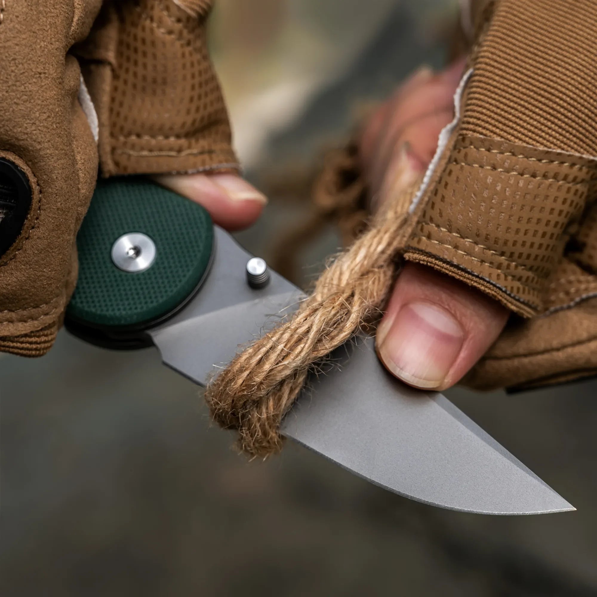 Gloved hands using folding knife with green handle to cut thick rope outdoors
