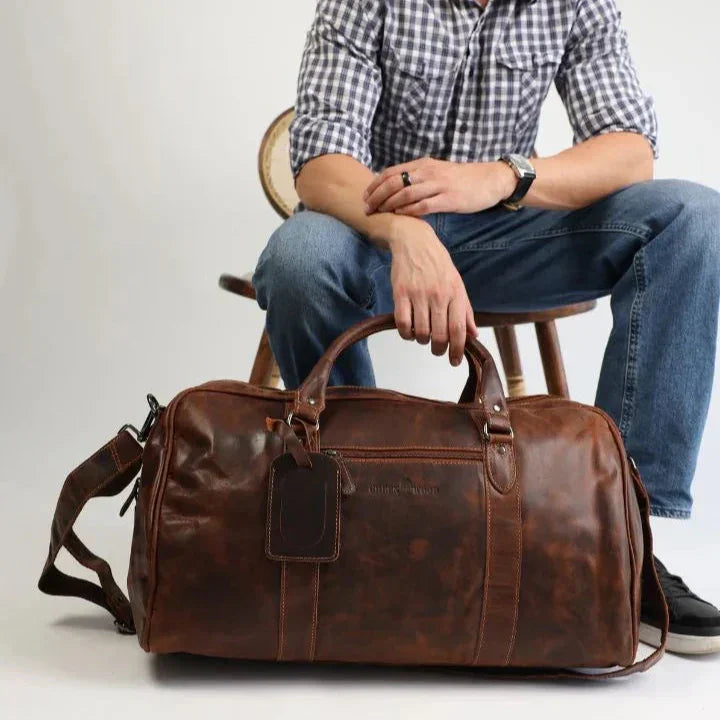 Man sitting with a brown leather duffel bag on the floor, casual travel style