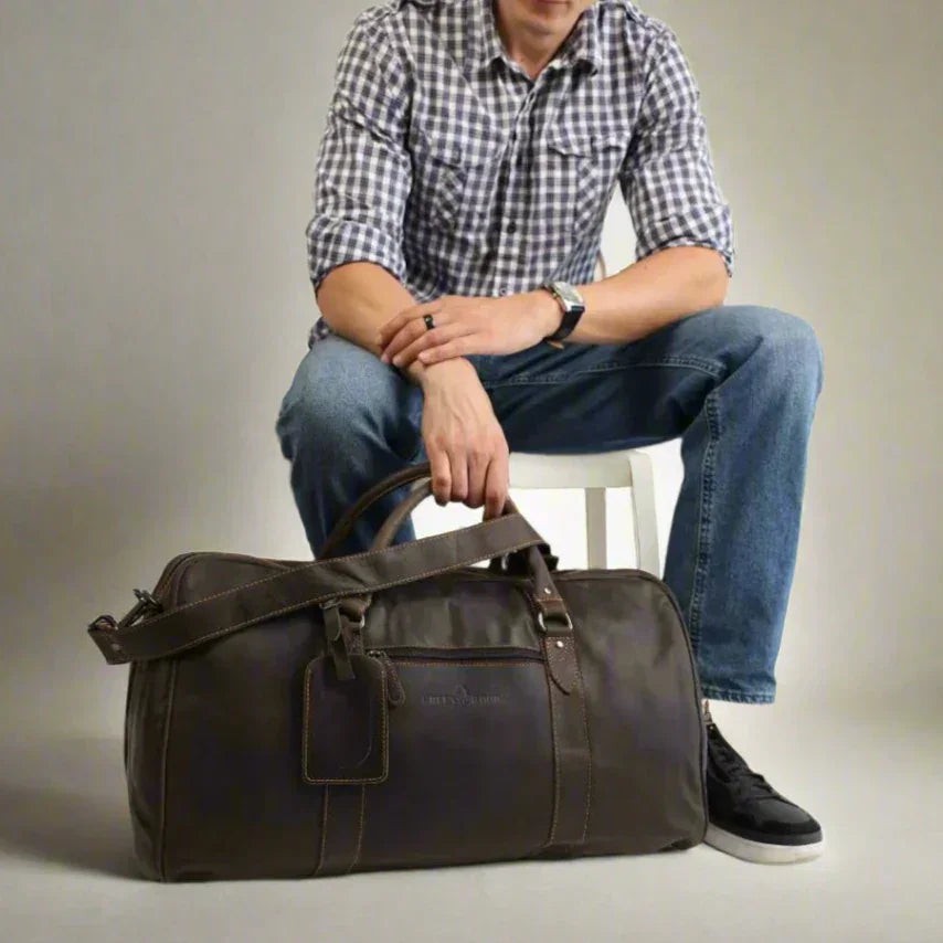Man in plaid shirt and jeans sitting with large brown leather duffel bag