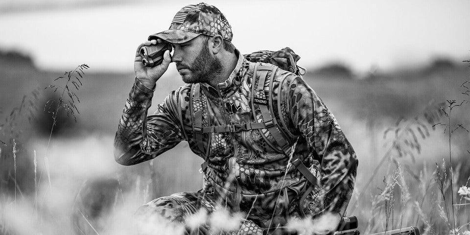 Hunter in camouflage gear with backpack using a rangefinder in tall grass field