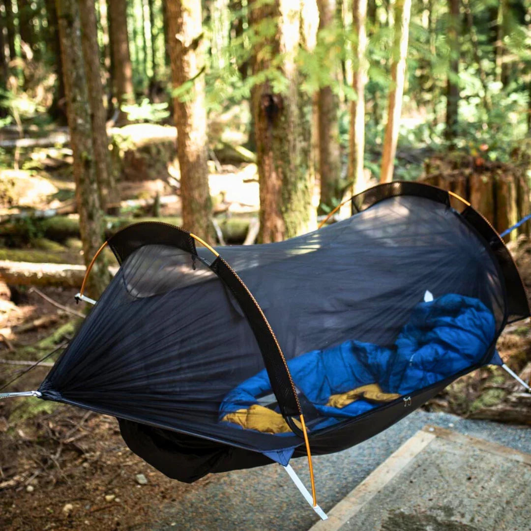 Mesh camping hammock with blue sleeping bag set up in a forested outdoor setting