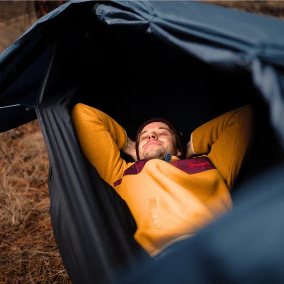Man relaxing in a camping hammock tent outdoors, wearing a yellow fleece jacket