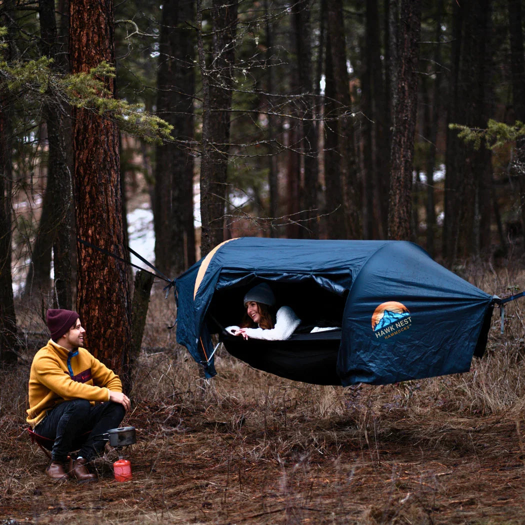 Two people camping in a forest, using a suspended Hawk Nest tent hammock.