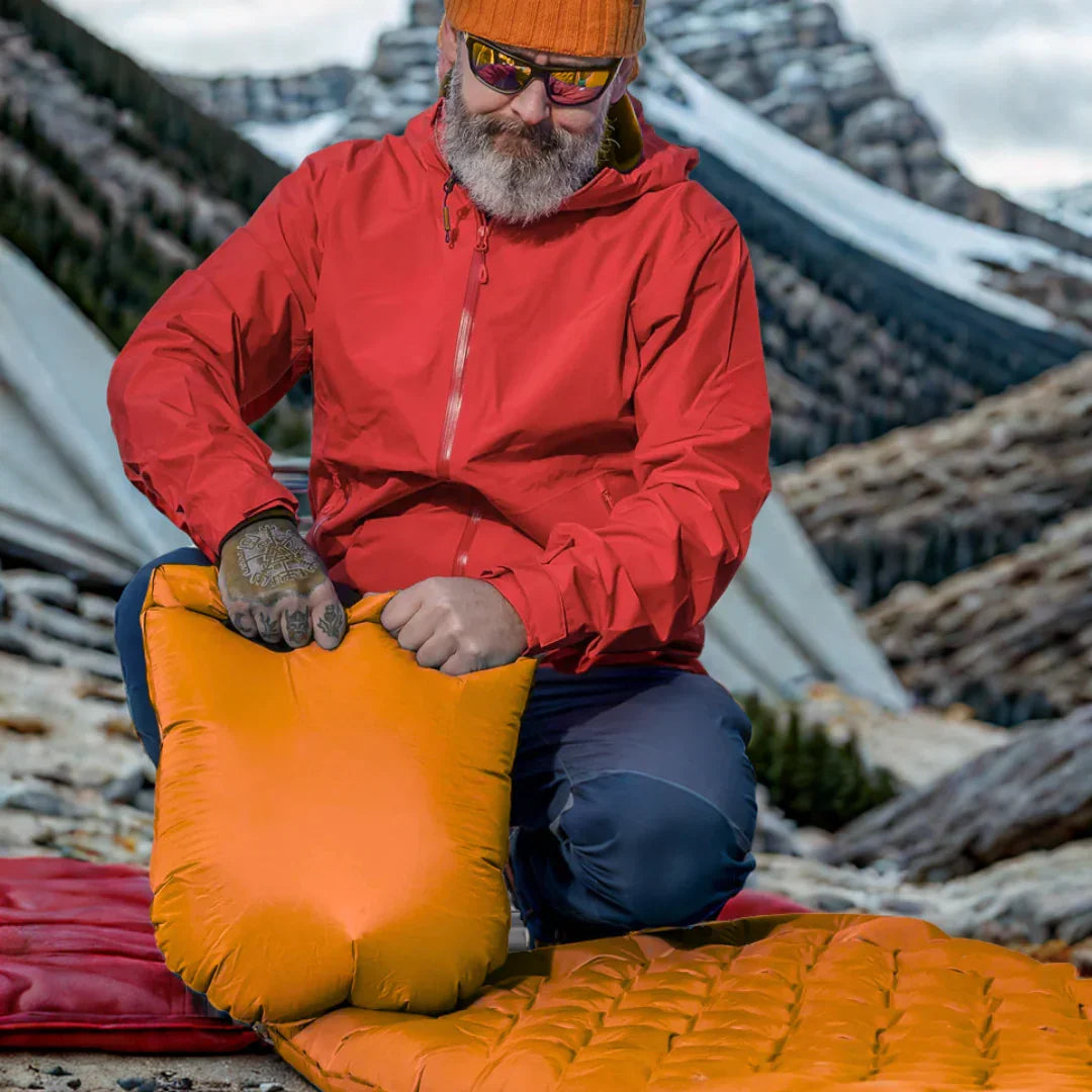 Man inflating orange camping sleeping pad outdoors in mountain setting