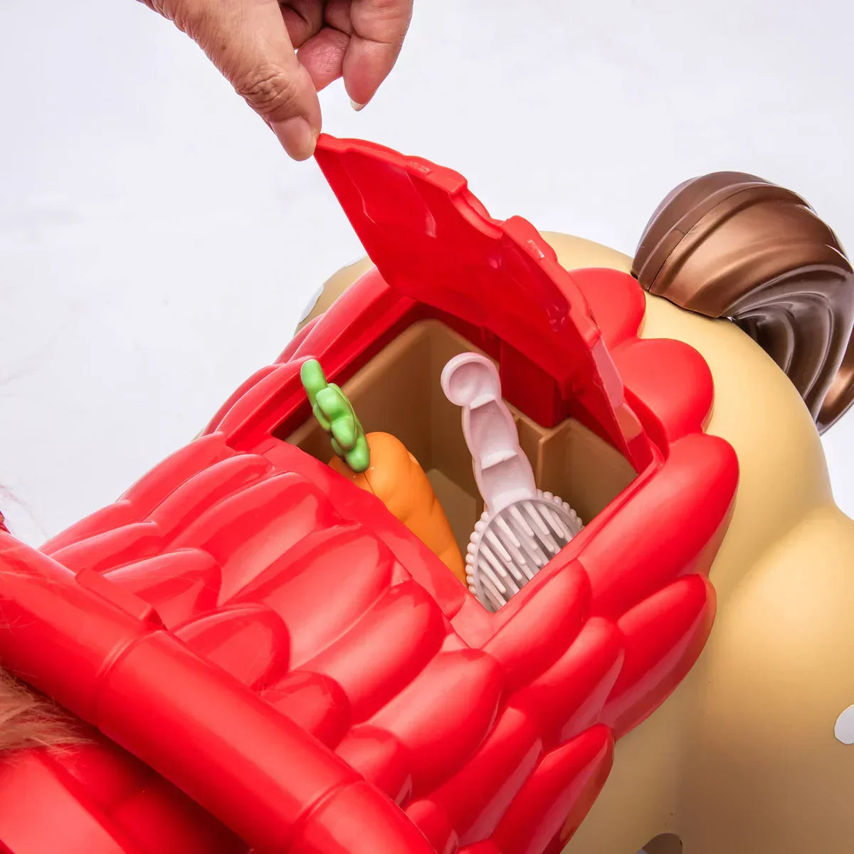 Hand opening red compartment on toy horse with toy carrot, brush, and bottle inside