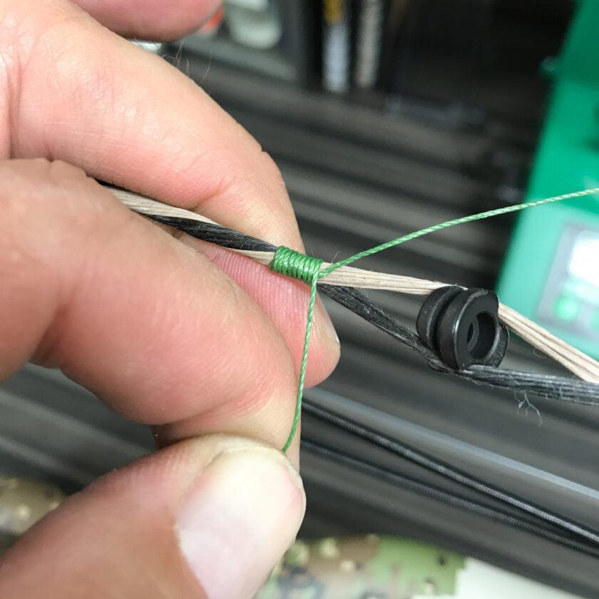Close-up of hand tying green thread on bowstring with peep sight, archery installation