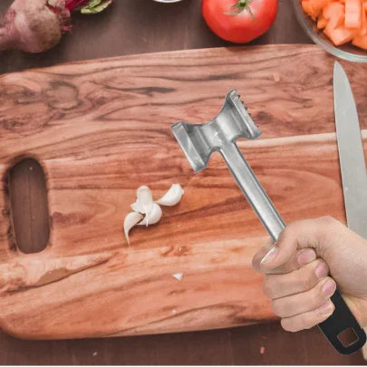 Hand holding metal meat tenderizer over wooden cutting board with garlic, knife, vegetables