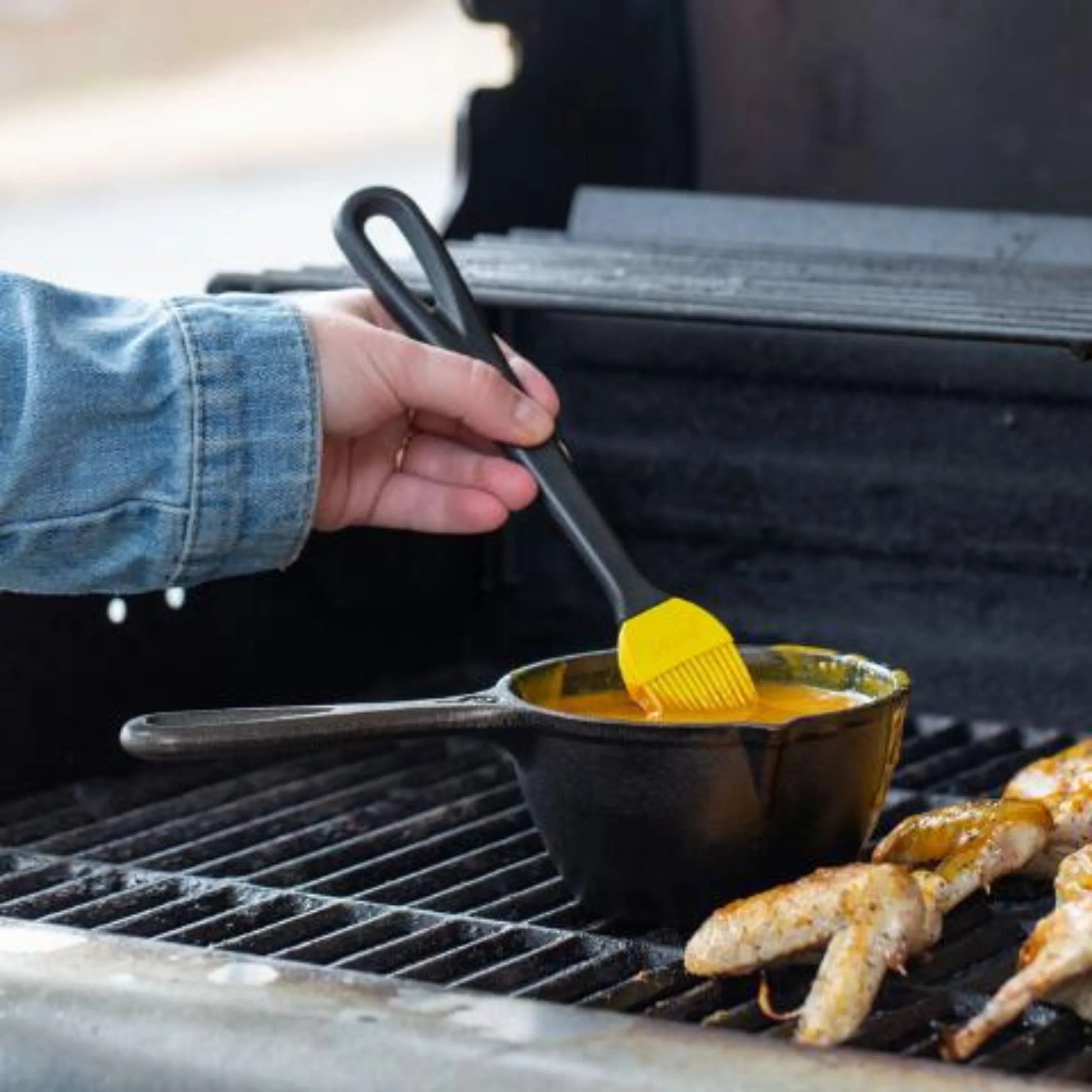 Hand using silicone brush with sauce in cast iron pot on outdoor grill, grilling chicken