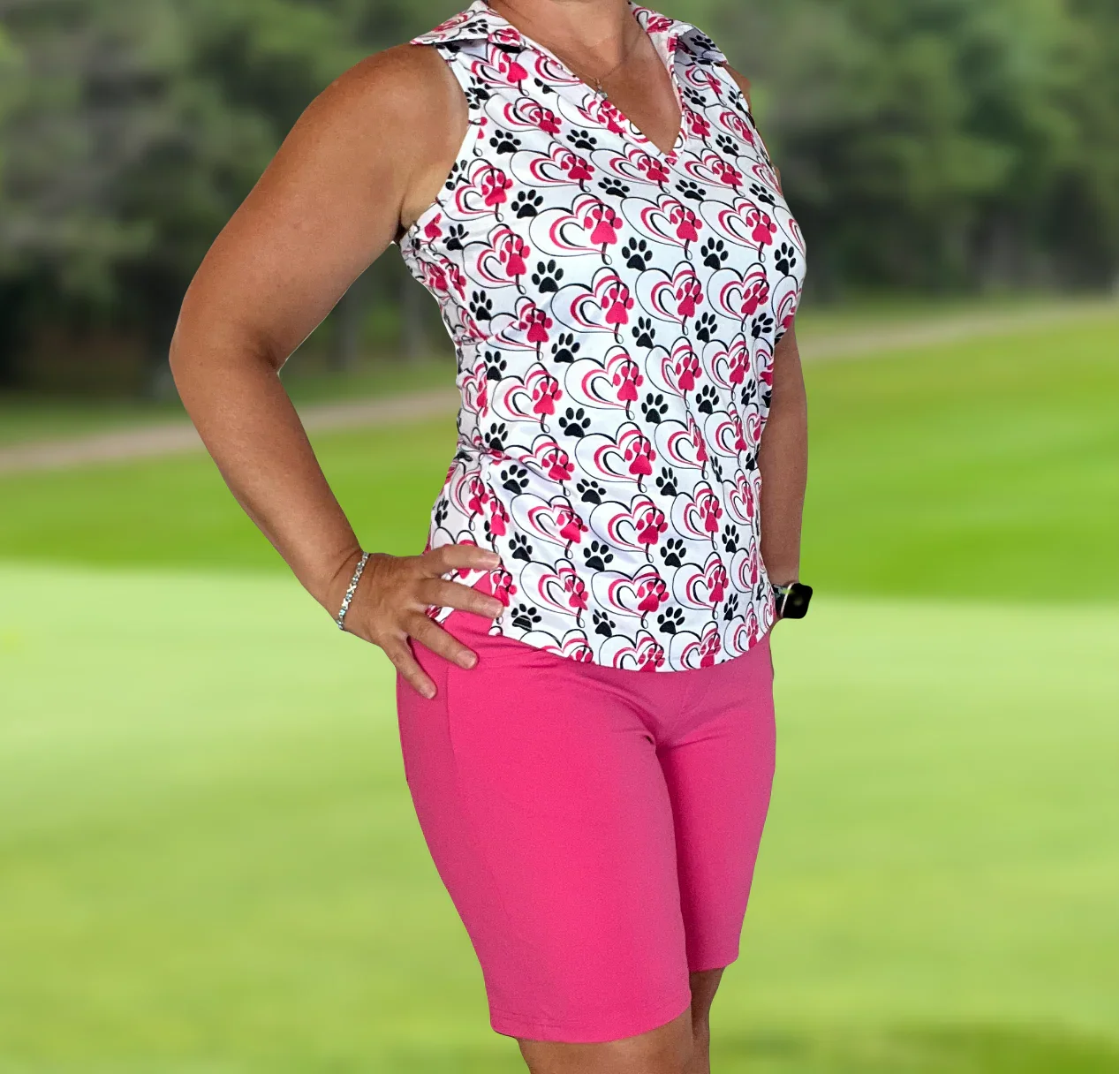 Woman wearing sleeveless heart and paw print golf polo with pink shorts stands on golf course